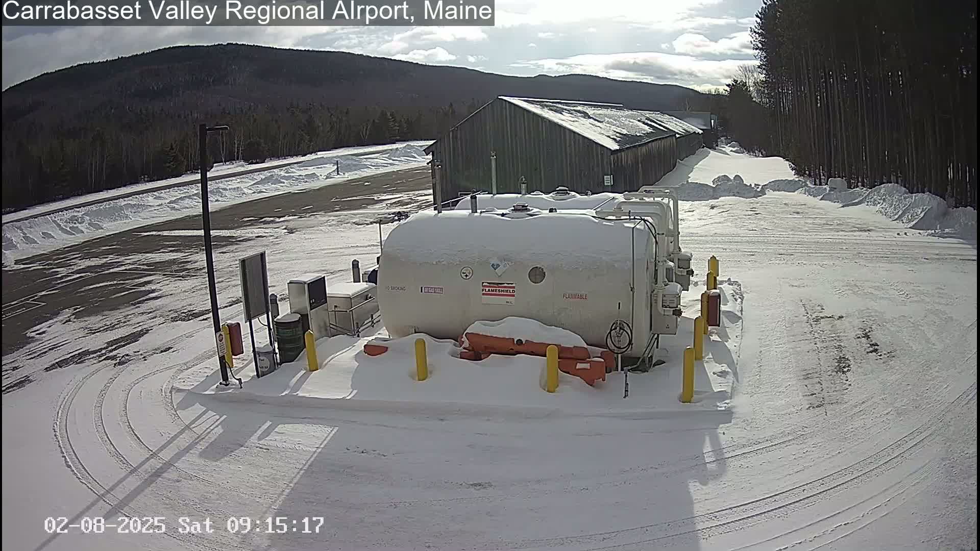 A snow-covered airport fueling station sits near a long, snow-covered building under a partly sunny sky.