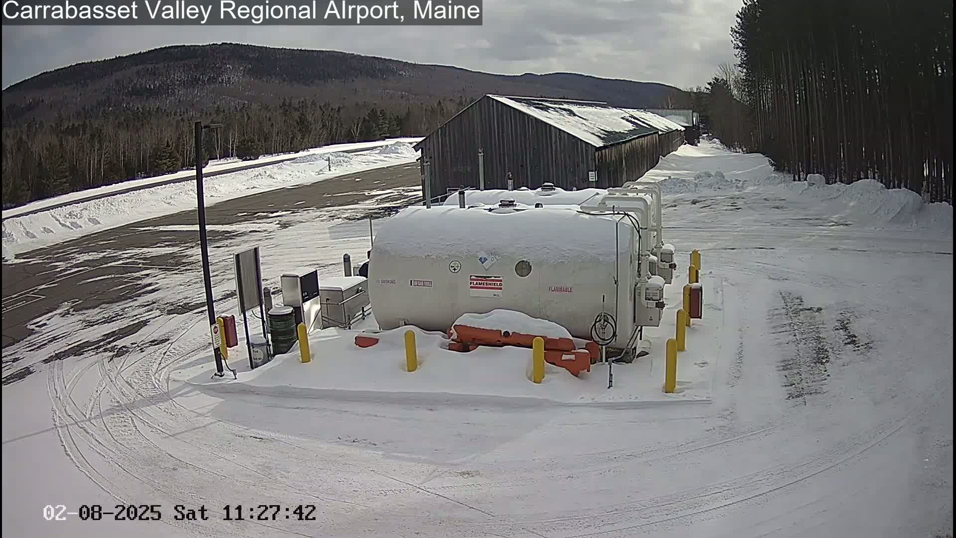 Several large white tanks covered in snow sit in a snow-covered parking lot near a long wooden building, with a mountain range visible in the background under a partly cloudy sky.