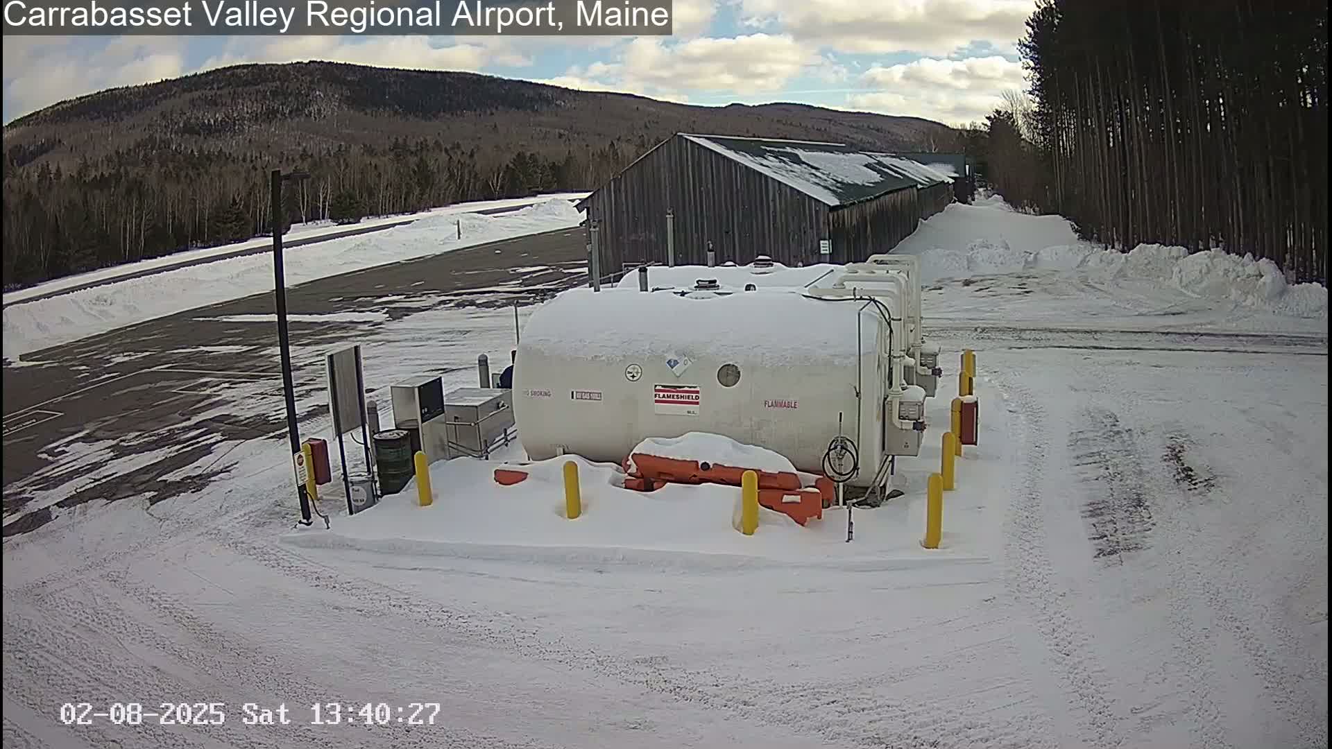 A snow-covered airport fuel farm sits in a snowy parking lot next to a long, gray building, with a mountain range visible in the background under a partly cloudy sky.