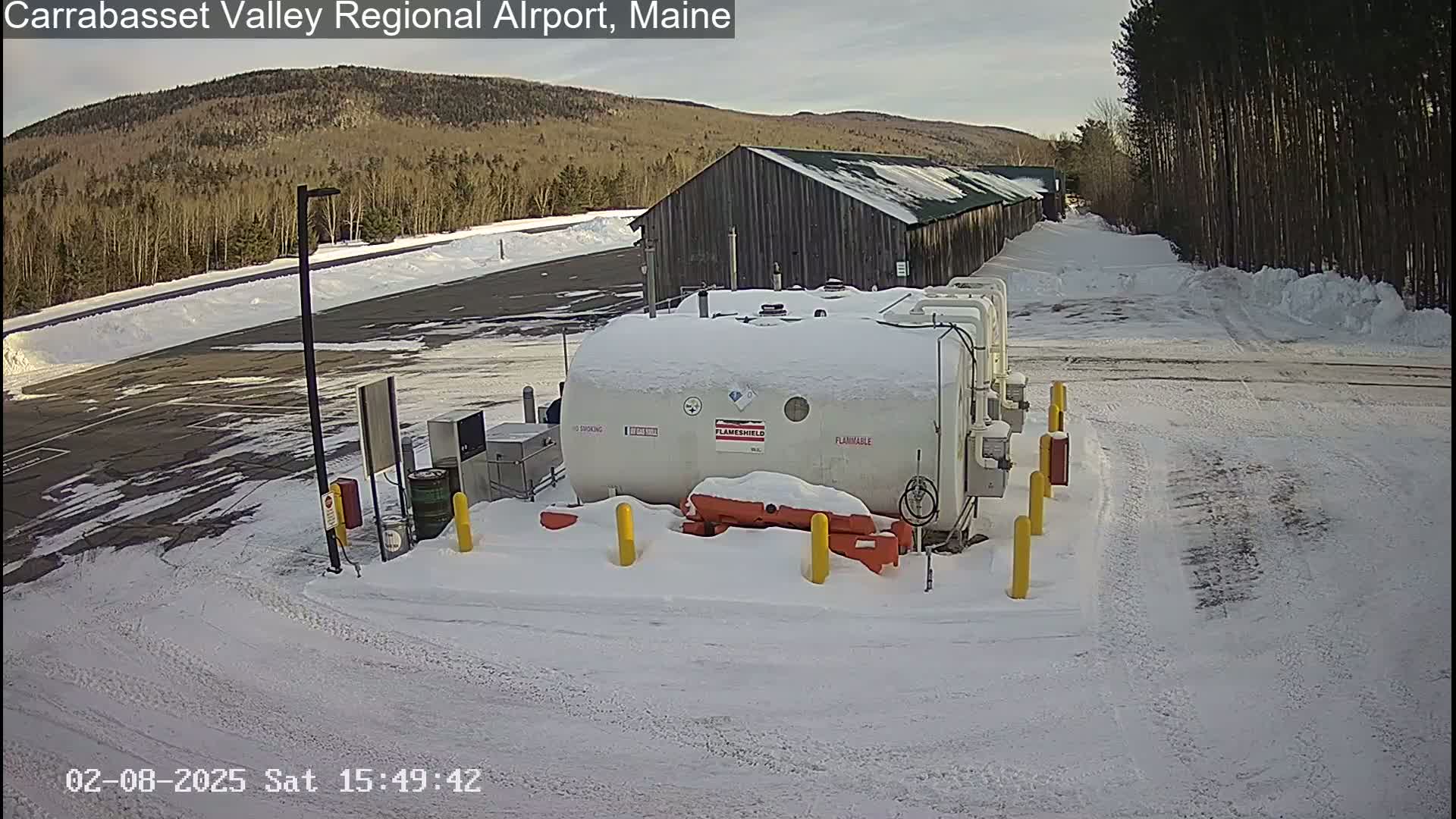 A snow-covered fuel tank farm sits in a mostly empty, snow-covered parking lot next to a long, dark building, with a background of trees and a distant mountain range under a mostly clear sky.