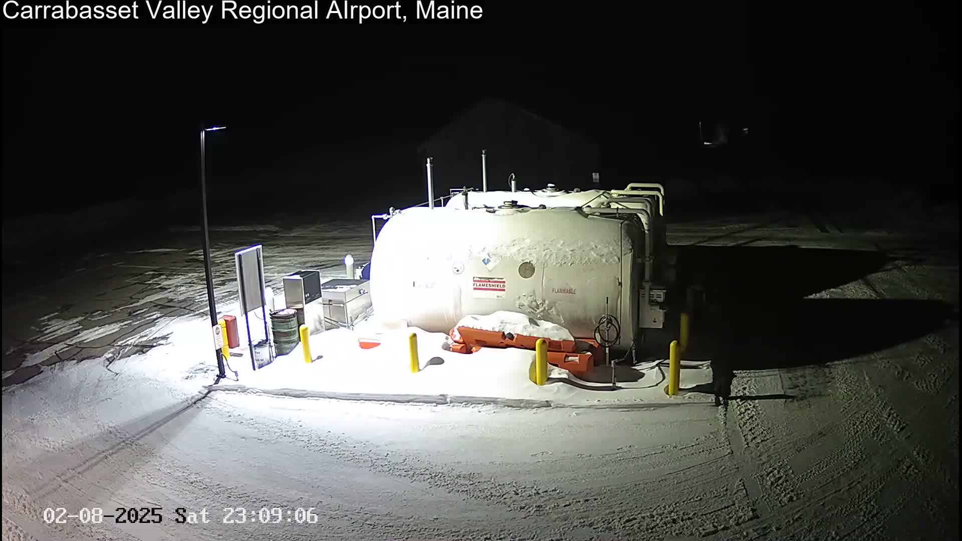 A snow-covered fuel tank sits in a snowy, nighttime airport parking lot illuminated by a single light pole.