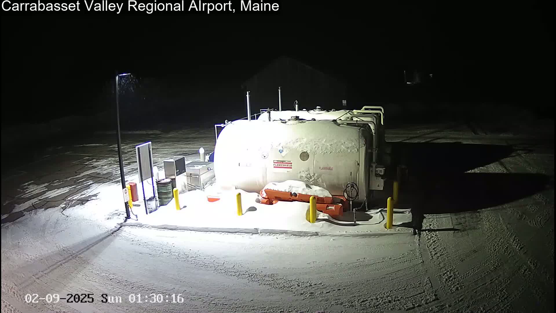 A snow-covered fuel storage tank sits in a dark, snowy, outdoor area at night, illuminated by artificial light.