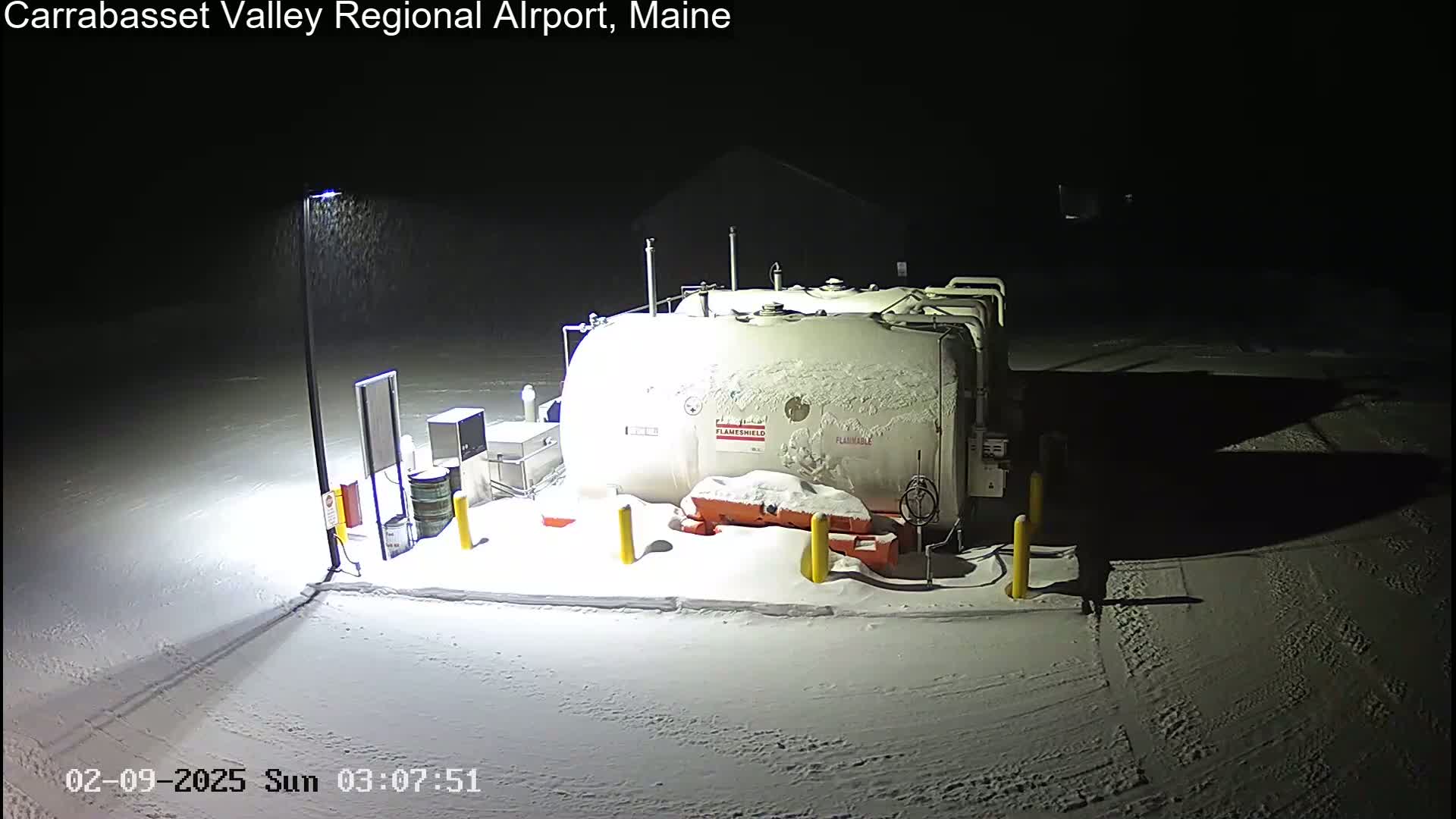 A large, snow-covered fuel tank sits in a snow-covered area at night under light snowfall.