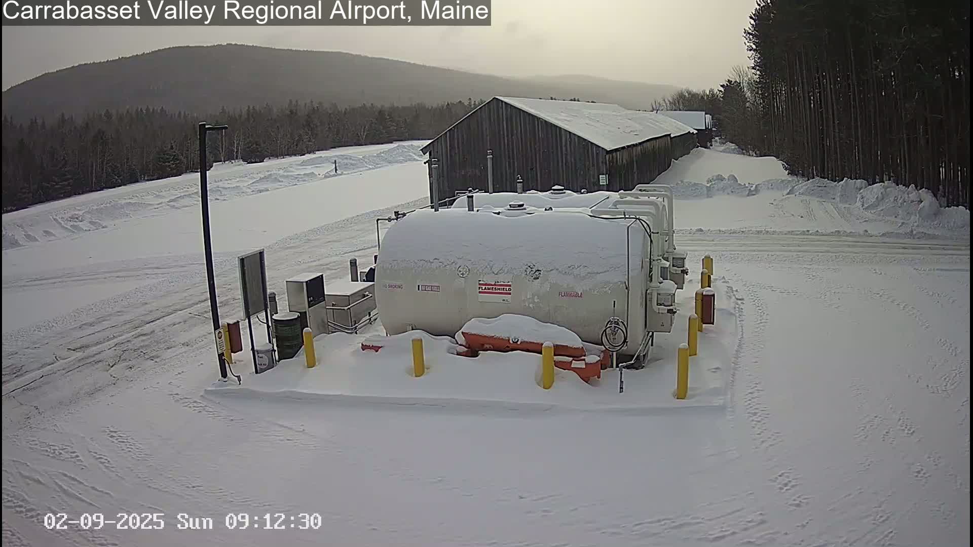 A snow-covered fuel storage facility sits near a long, snow-covered building, with a background of snow-covered trees and distant hills under an overcast sky.