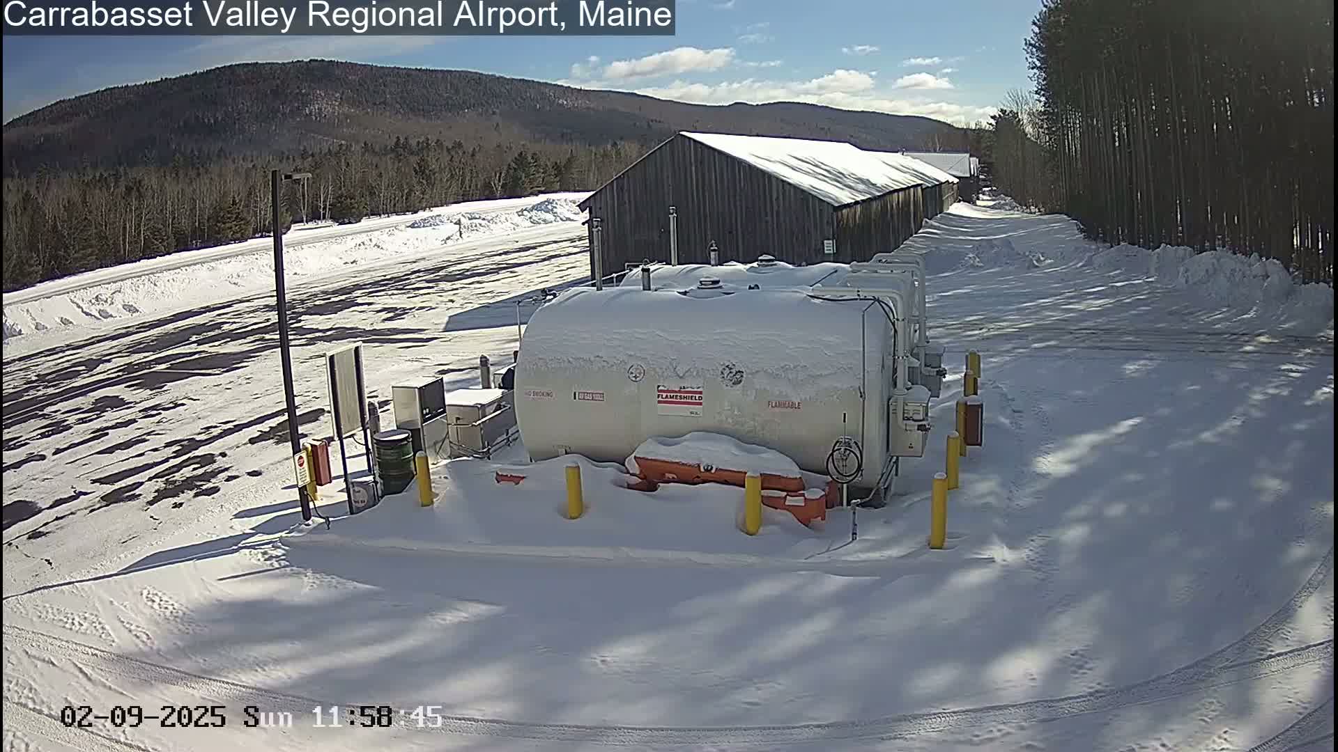 A snow-covered fuel storage area with a large tank, a nearby building, and trees in the background under a sunny sky.