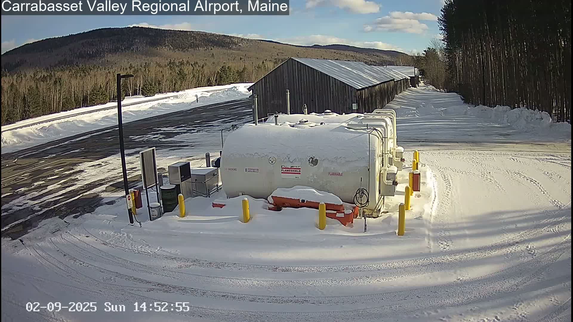A snow-covered airport fuel depot sits near a long wooden building under a sunny sky.