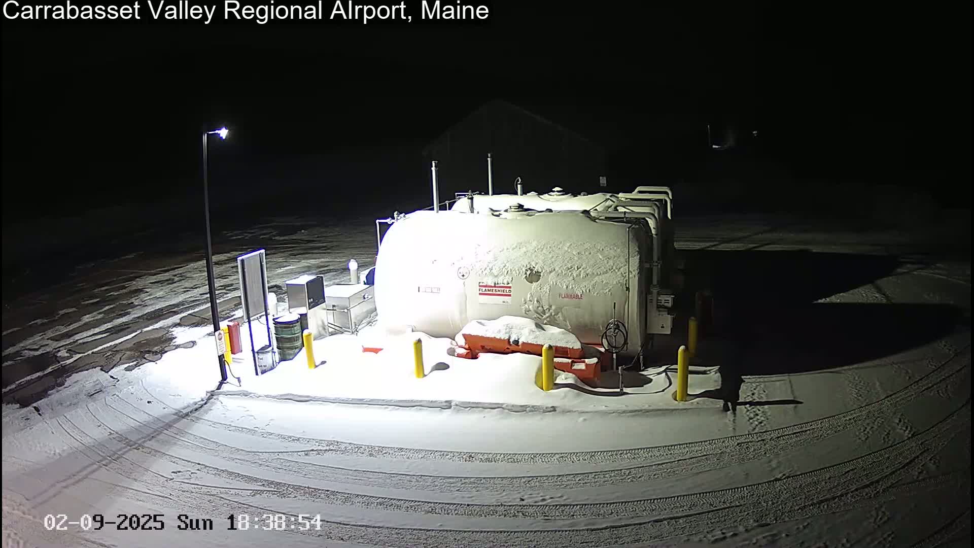 A snow-covered fuel tank and associated equipment are illuminated at night in a snowy area.