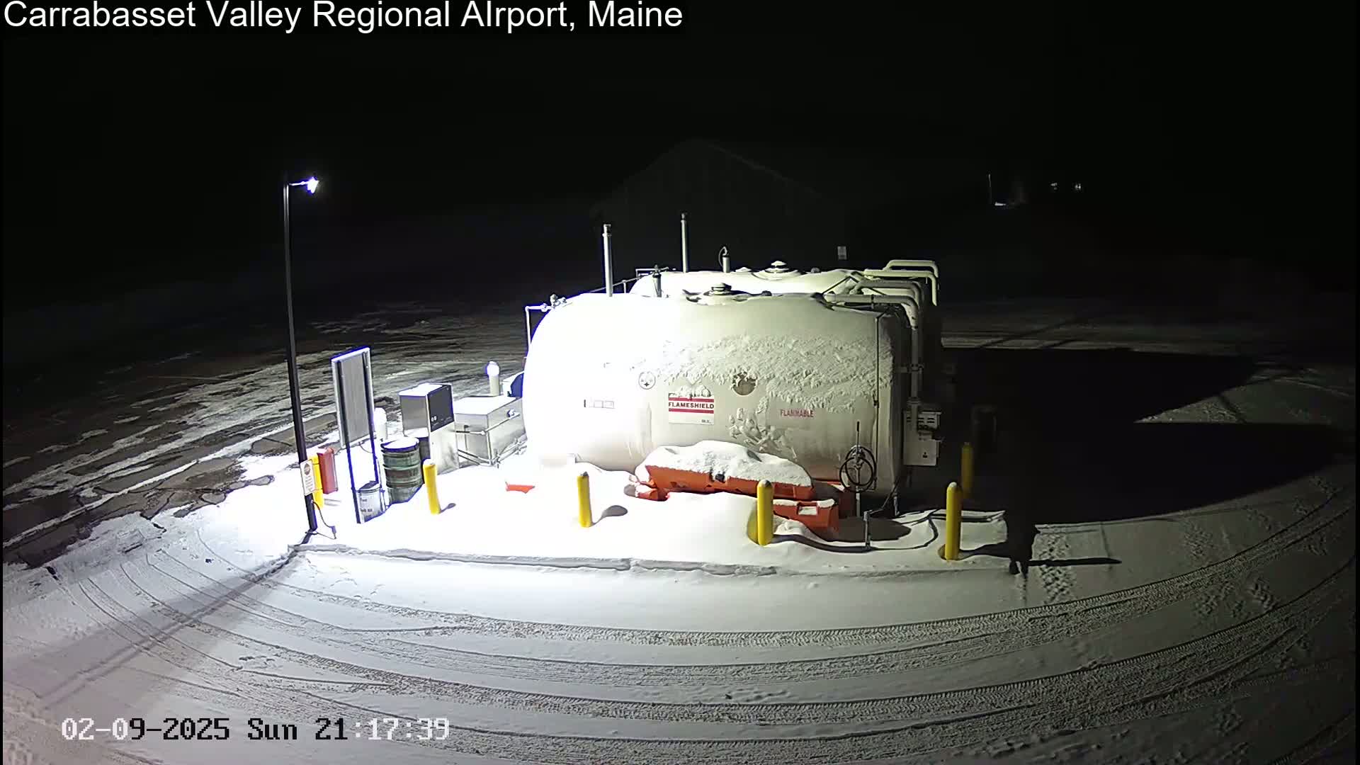 A snow-covered fuel tank sits in a snow-covered parking lot at night, illuminated by a single lamp.