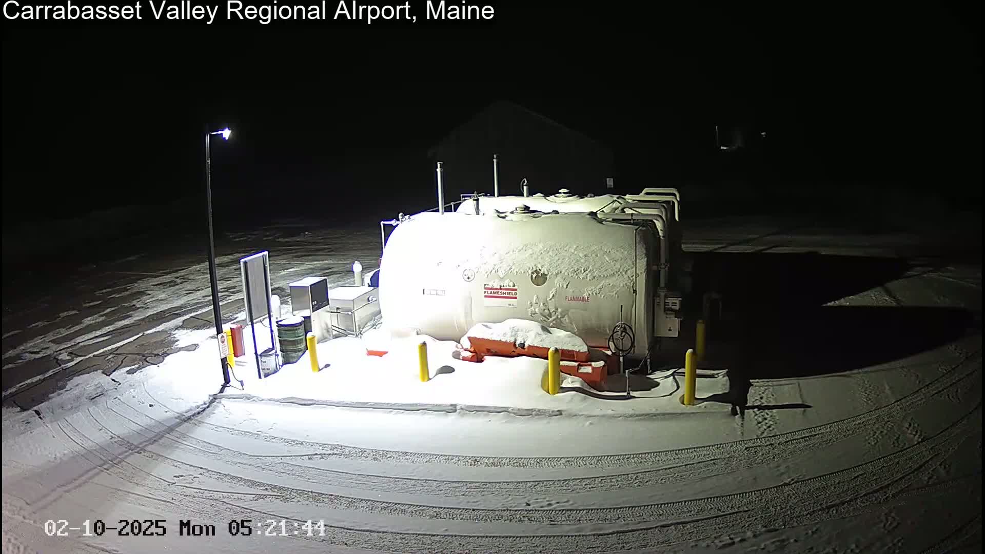 A snow-covered fuel tank and surrounding equipment are illuminated at night in a snowy parking lot.