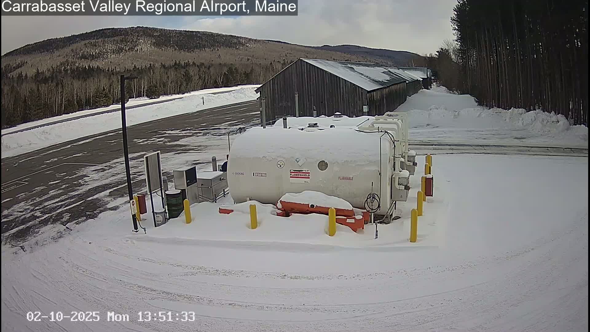 A snow-covered airport fueling station sits in a snowy lot next to a long, dark building, with a wooded mountain in the background under a cloudy sky.