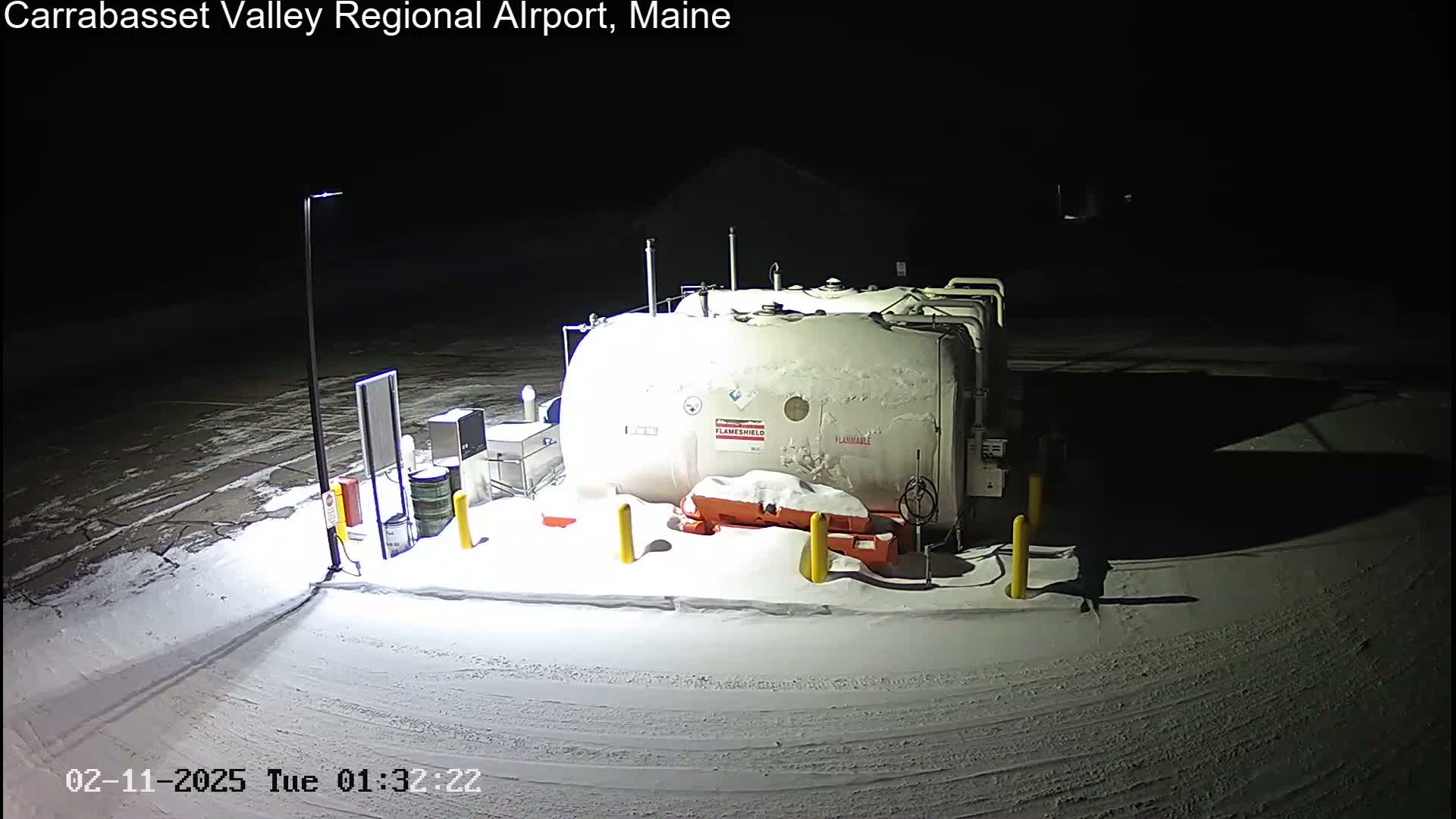 A large, snow-covered fuel tank sits in a snow-covered airport lot at night.