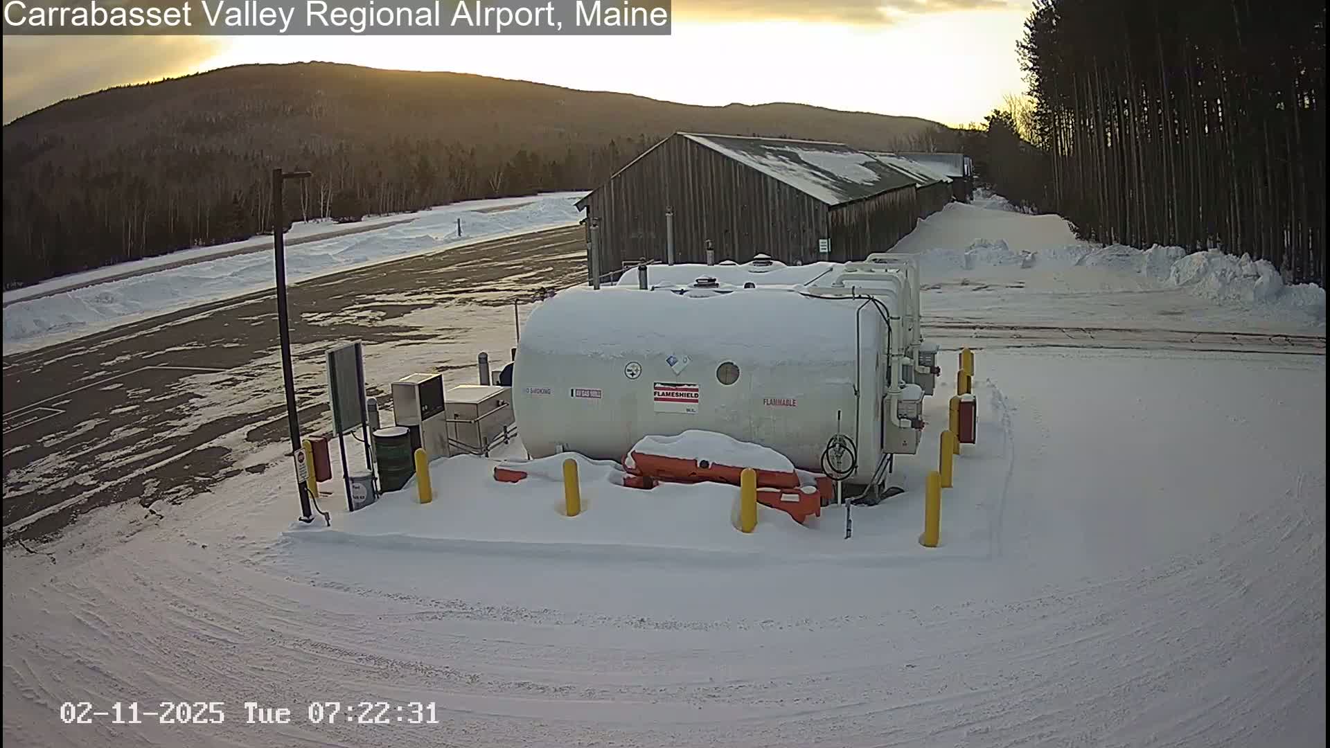 A snow-covered fuel storage tank sits in a snowy airport parking lot near a long building, with a mountain range visible in the background under a partly cloudy sky.