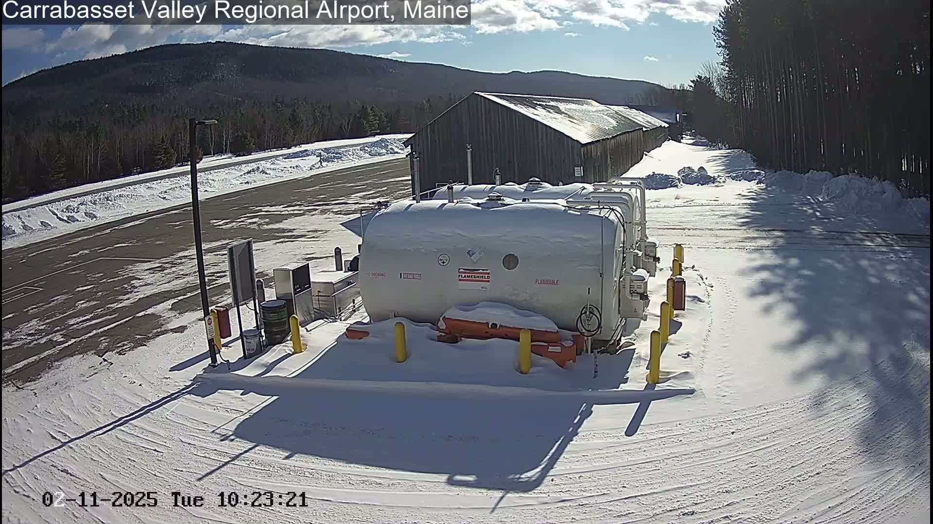 Under a partly sunny sky, a snow-covered airport area features several large white fuel tanks near a long wooden building, with tire tracks in the snow surrounding them.