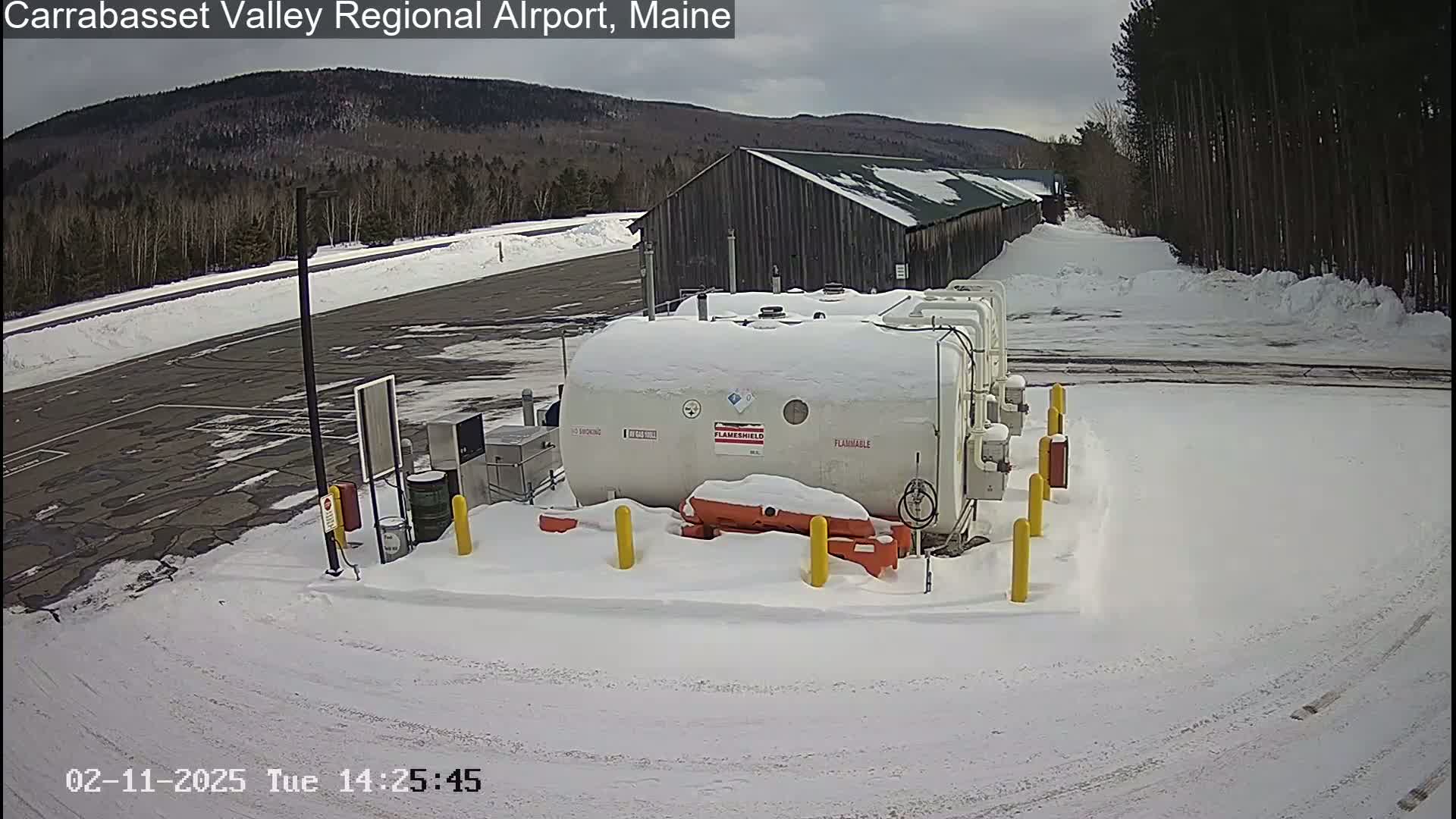 A snow-covered airport fueling station with a large white tank sits in a snow-covered parking lot, with a wooded area and mountains in the background under a cloudy sky.