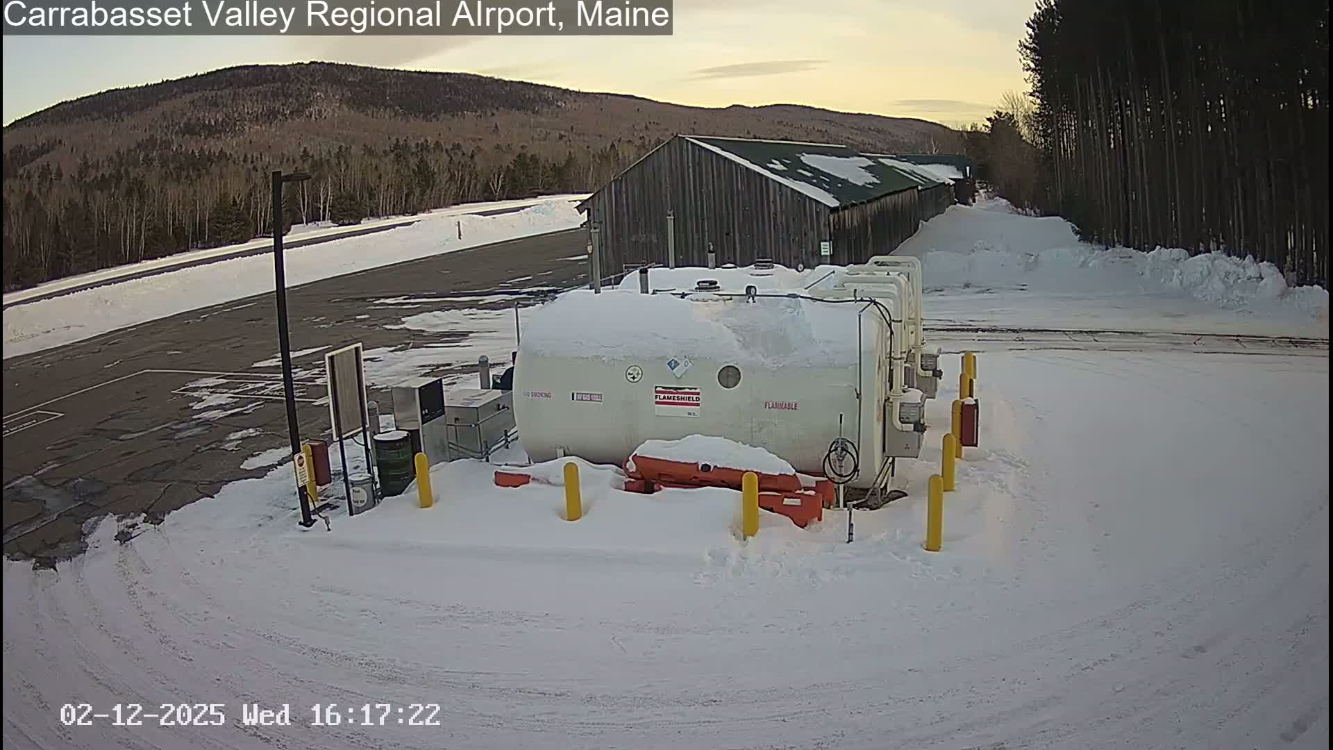 A snow-covered airport fuel storage area is situated near a long, dark building, with a background of snow-covered hills and trees under a twilight sky.