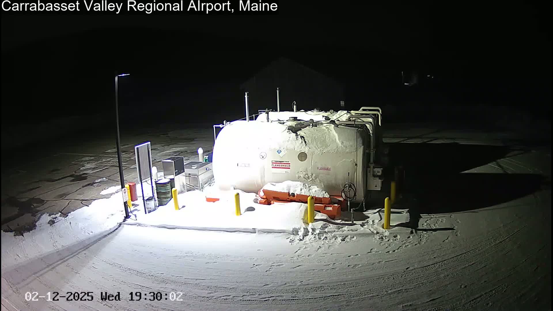 A large, snow-covered fuel tank sits in a snow-covered parking lot at night, illuminated by a single light pole.