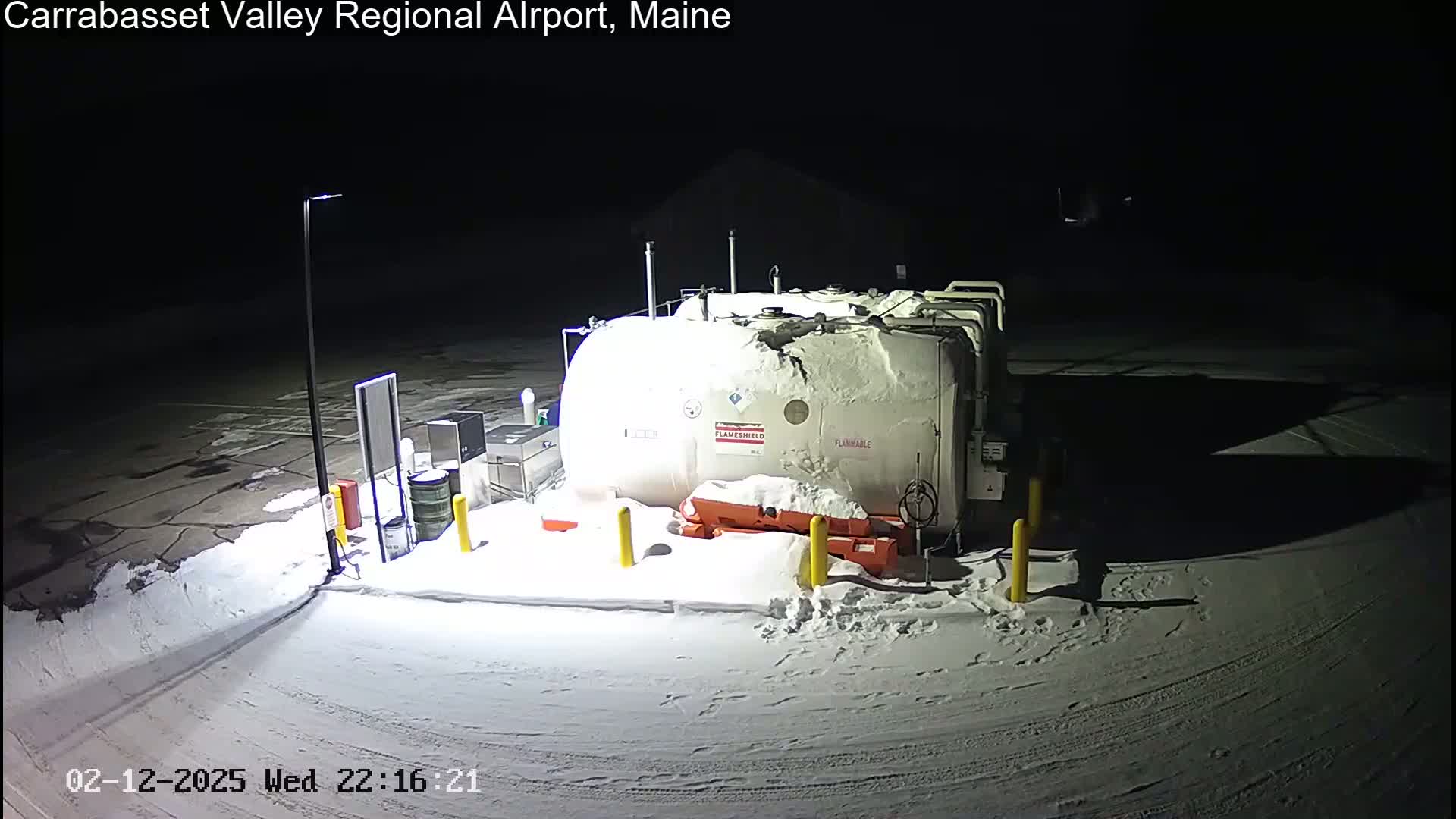 A large, snow-covered fuel tank sits in a snow-covered parking lot at night, illuminated by artificial light.