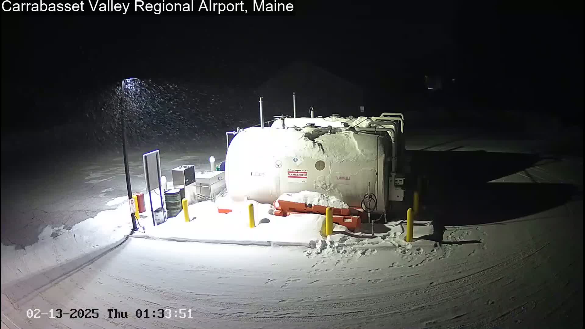 A large, snow-covered fuel tank sits in a snow-covered parking lot at night under light snowfall.