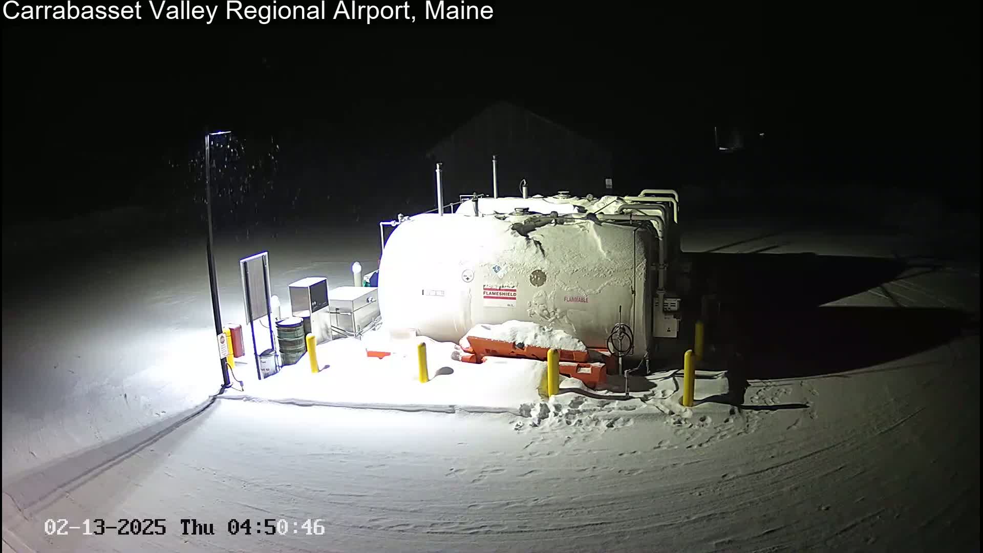 A large, snow-covered fuel tank sits in a snowy, nighttime airport setting under light snowfall.