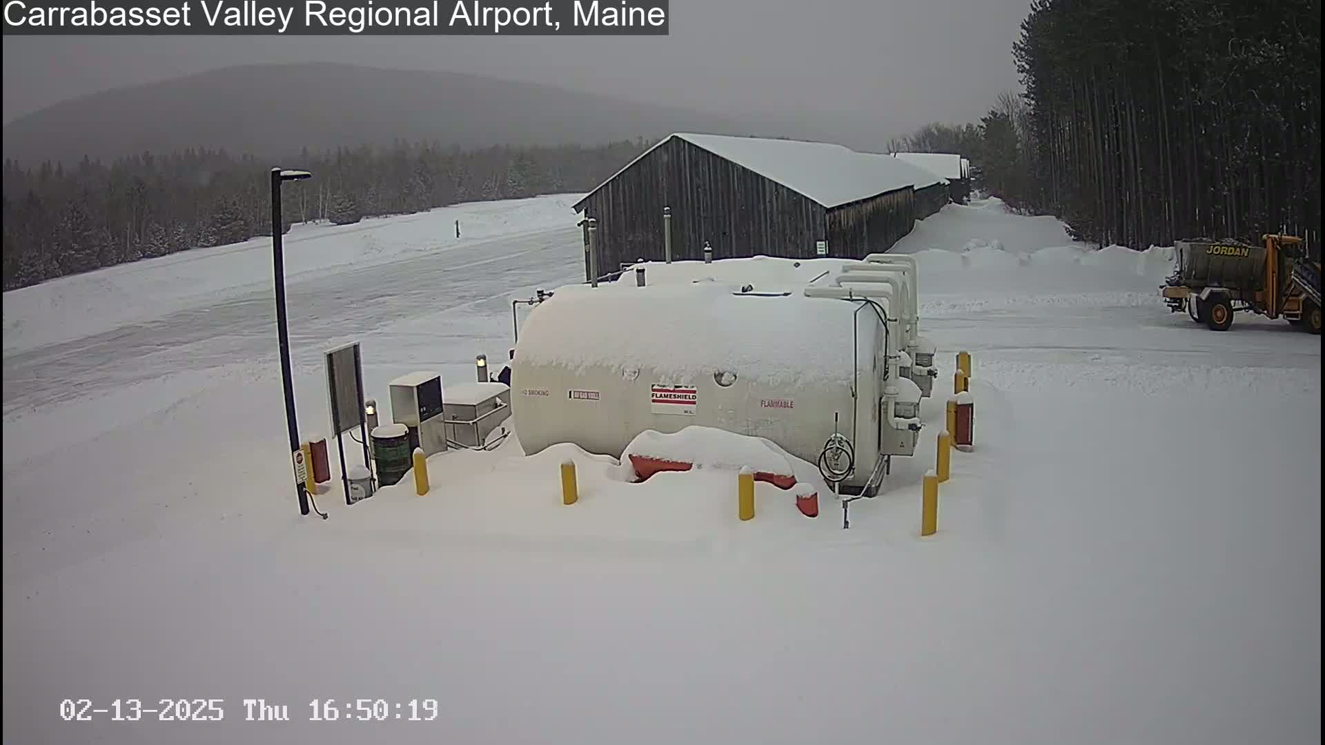 A snow-covered fuel tank and a nearby building are visible in a snowy landscape with a small yellow vehicle in the background.