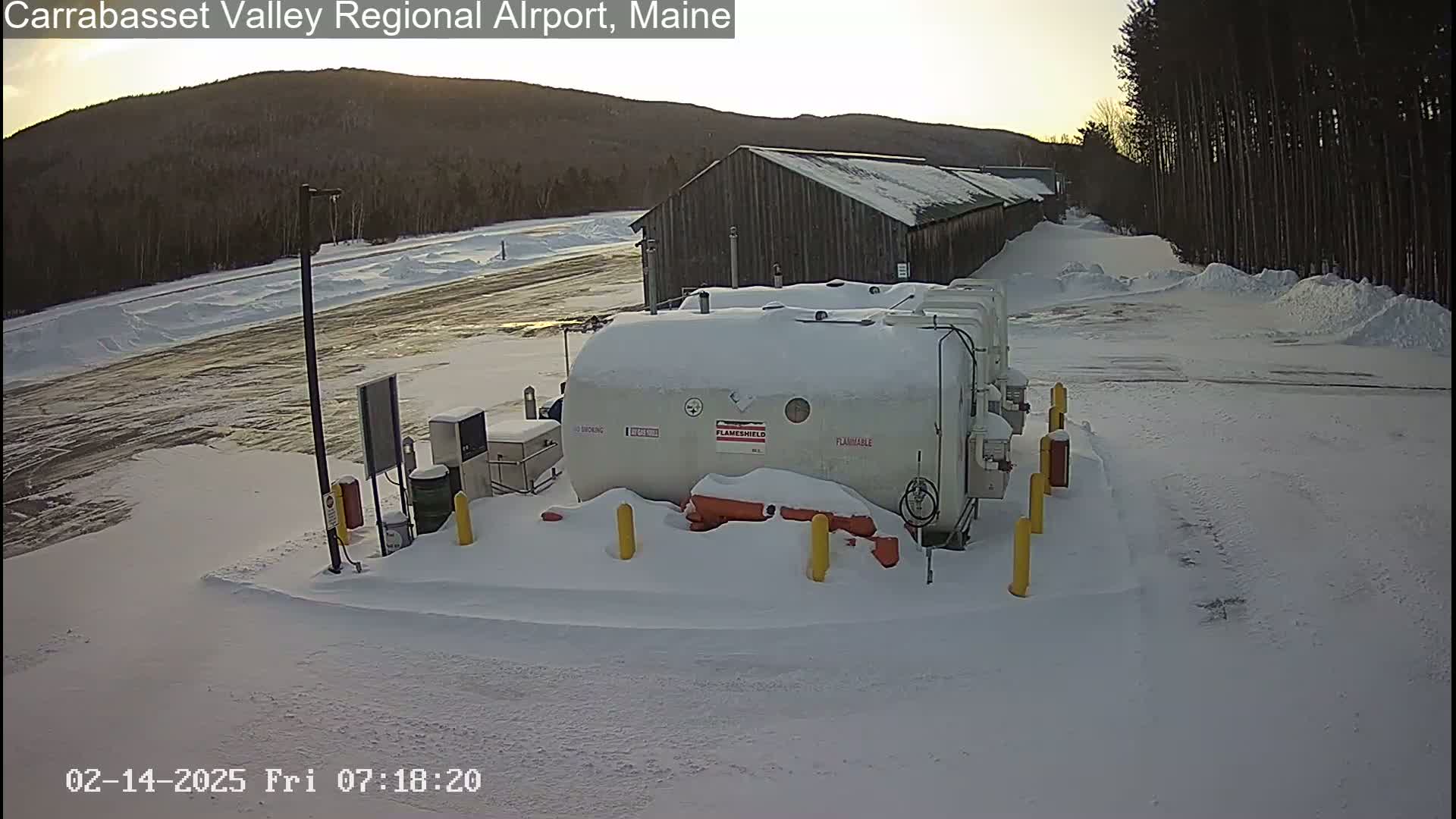 Several large, white, snow-covered fuel tanks sit in a snowy airport lot near a long, dark building under a partly sunny sky.