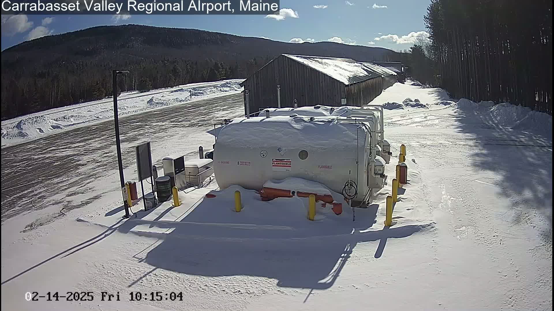 A snow-covered airport fuel storage area sits before a snow-covered, tree-lined landscape under a partly sunny sky.