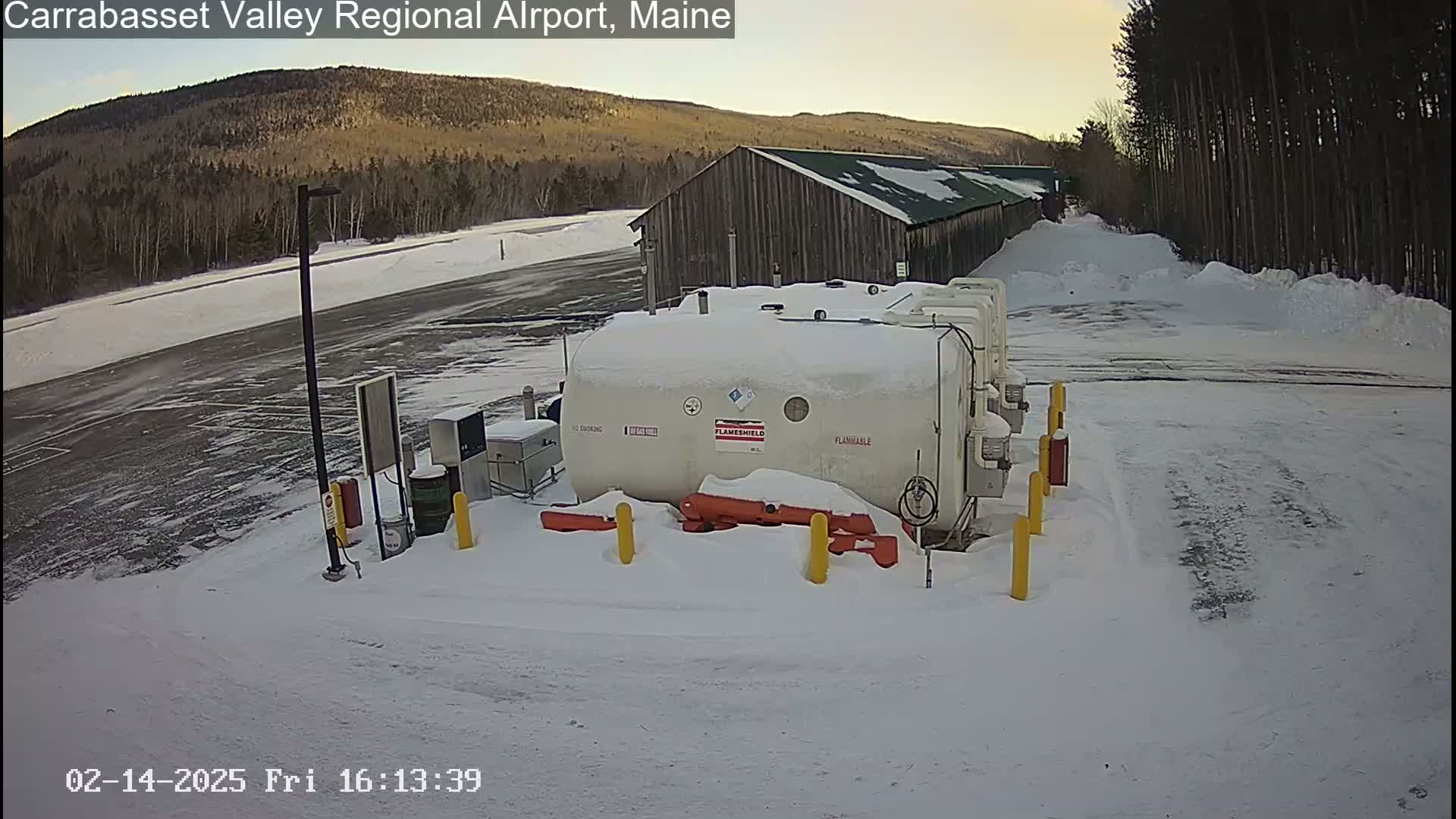 A large, white fuel tank sits in a snow-covered airport parking lot near a wooden building, with a background of a snow-covered landscape and distant mountains under a mostly clear sky.