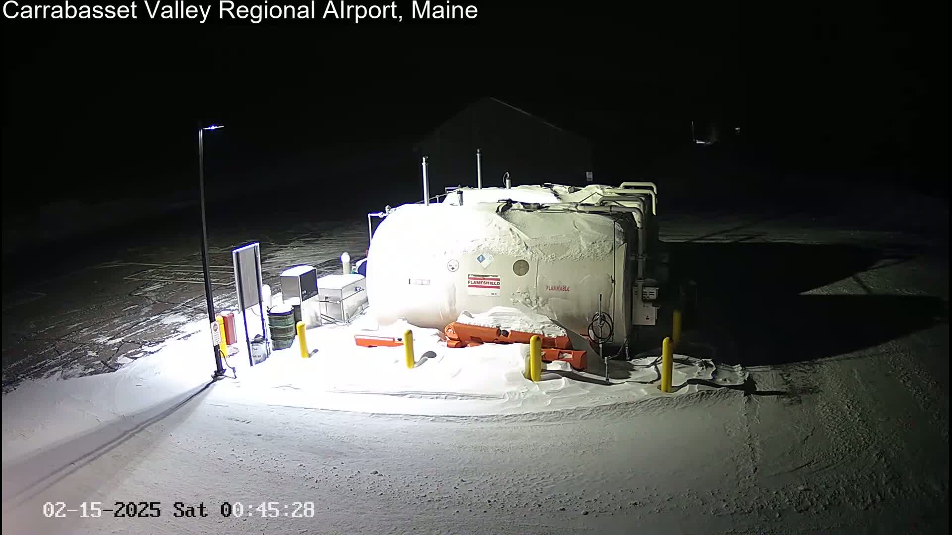 A large, snow-covered fuel tank sits in a snow-covered parking lot at night, illuminated by a single lamppost.