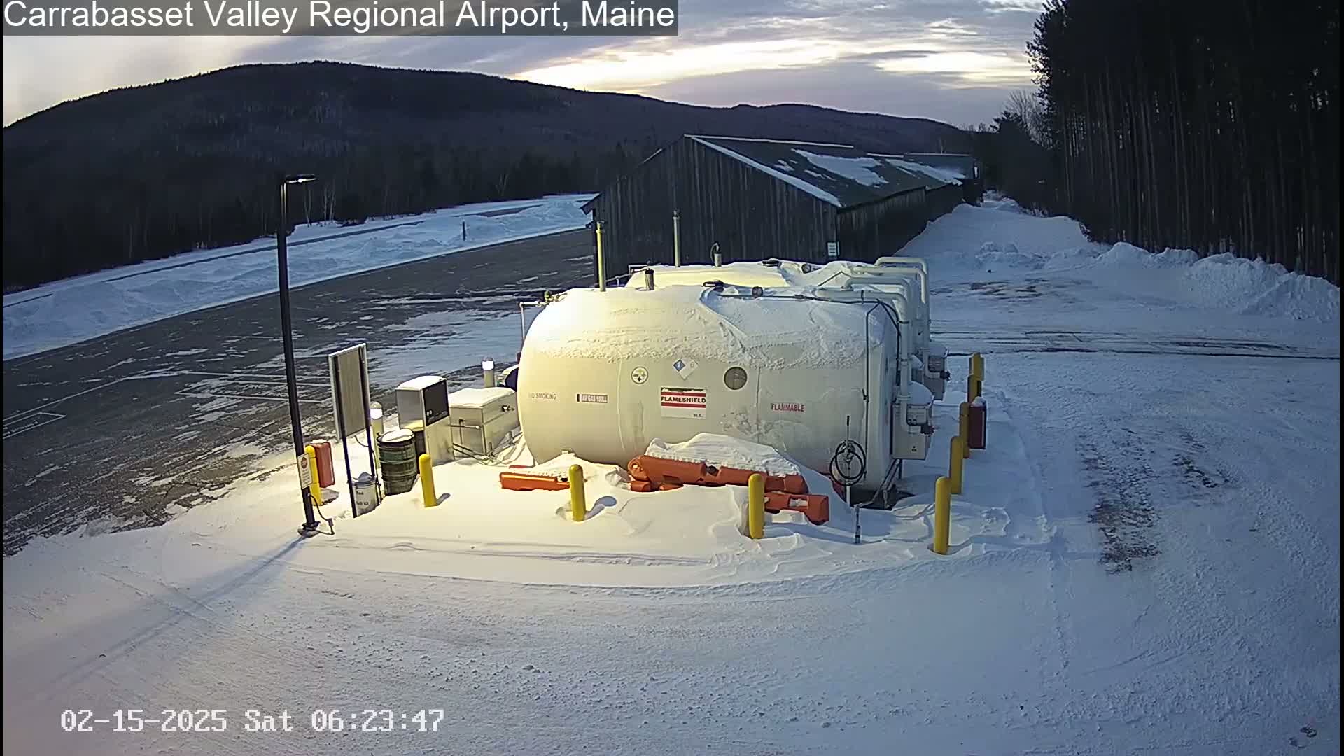 A snow-covered airport tarmac with several large, cylindrical tanks and a building in the background under a partly cloudy sky.
