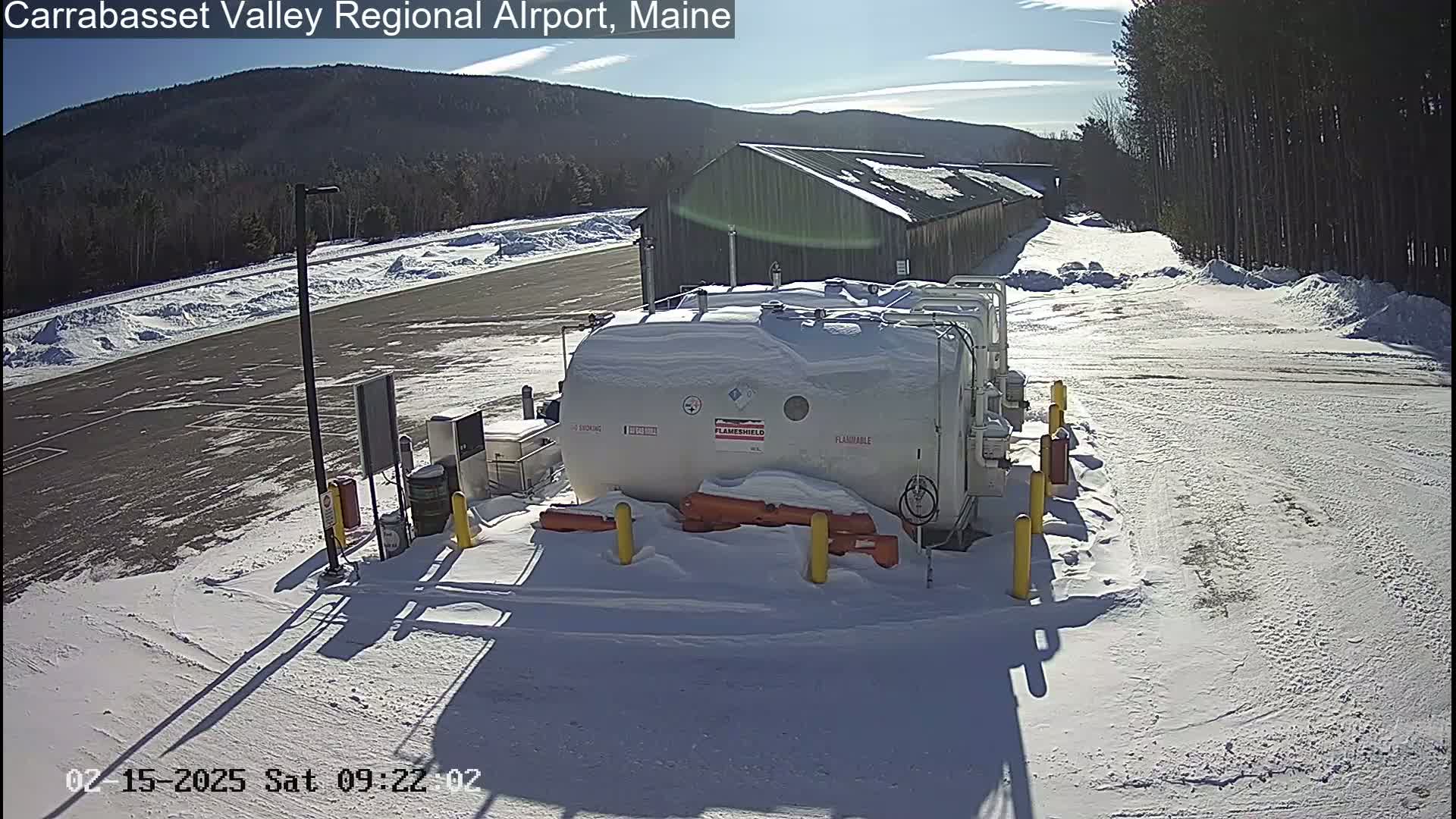 A snow-covered airport fueling station sits near a long building under a sunny sky.