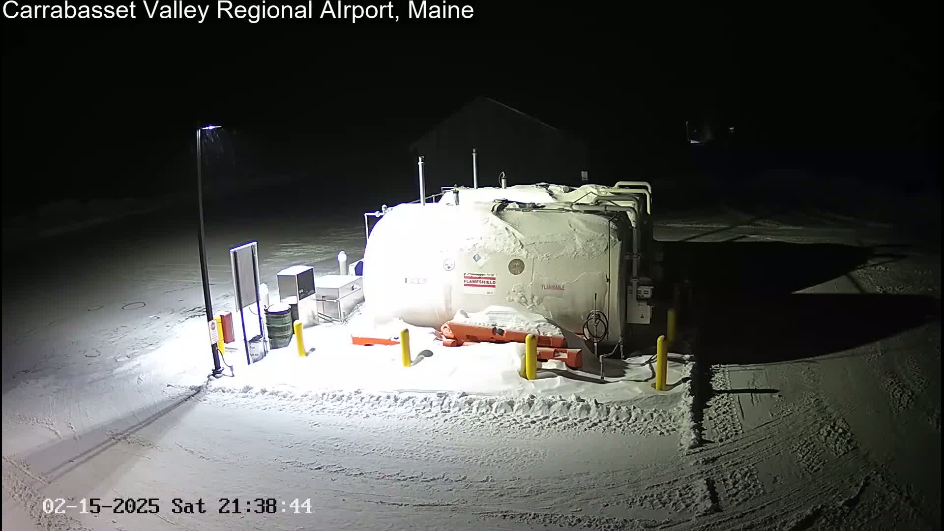 A large, snow-covered fuel tank sits in a snow-covered area at night, illuminated by a single lamppost.