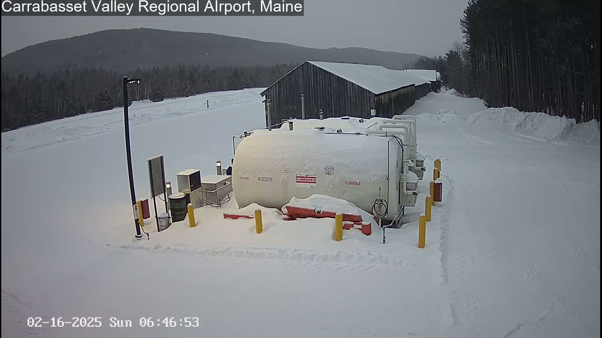 A large, snow-covered fuel tank sits in a snowy area near a dark-colored building, with a background of snow-covered trees and mountains under an overcast sky.