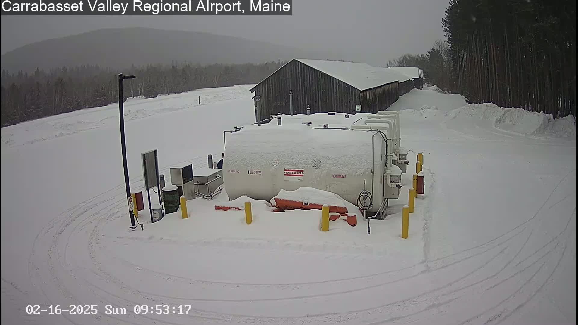 A snow-covered fuel tank sits in a snowy, outdoor area next to a long, gray building, with tire tracks visible in the snow.