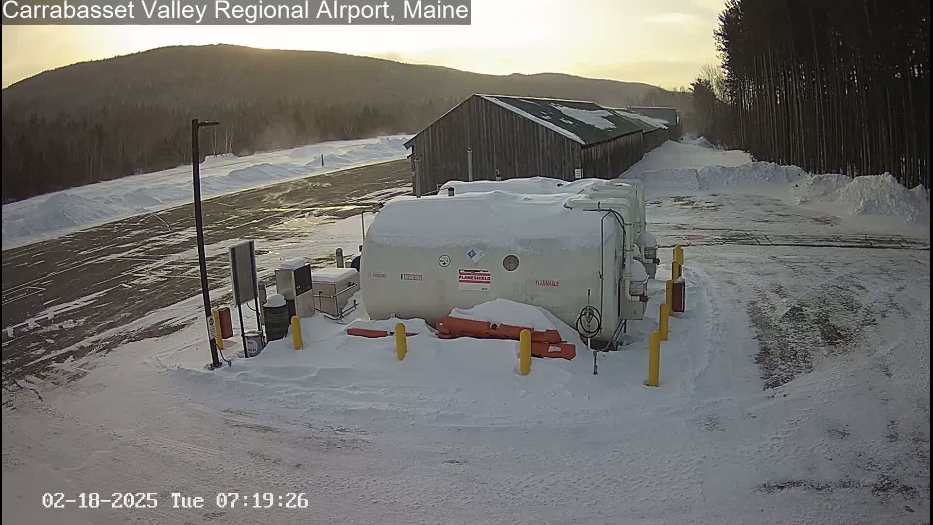 A snow-covered airport fuel depot with a large, snow-covered storage tank sits next to a long, wooden building in a snowy landscape.