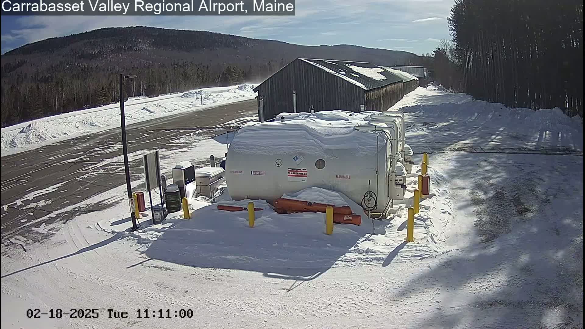 A snowy airport fuel farm sits next to a long, snow-covered building under a sunny sky.