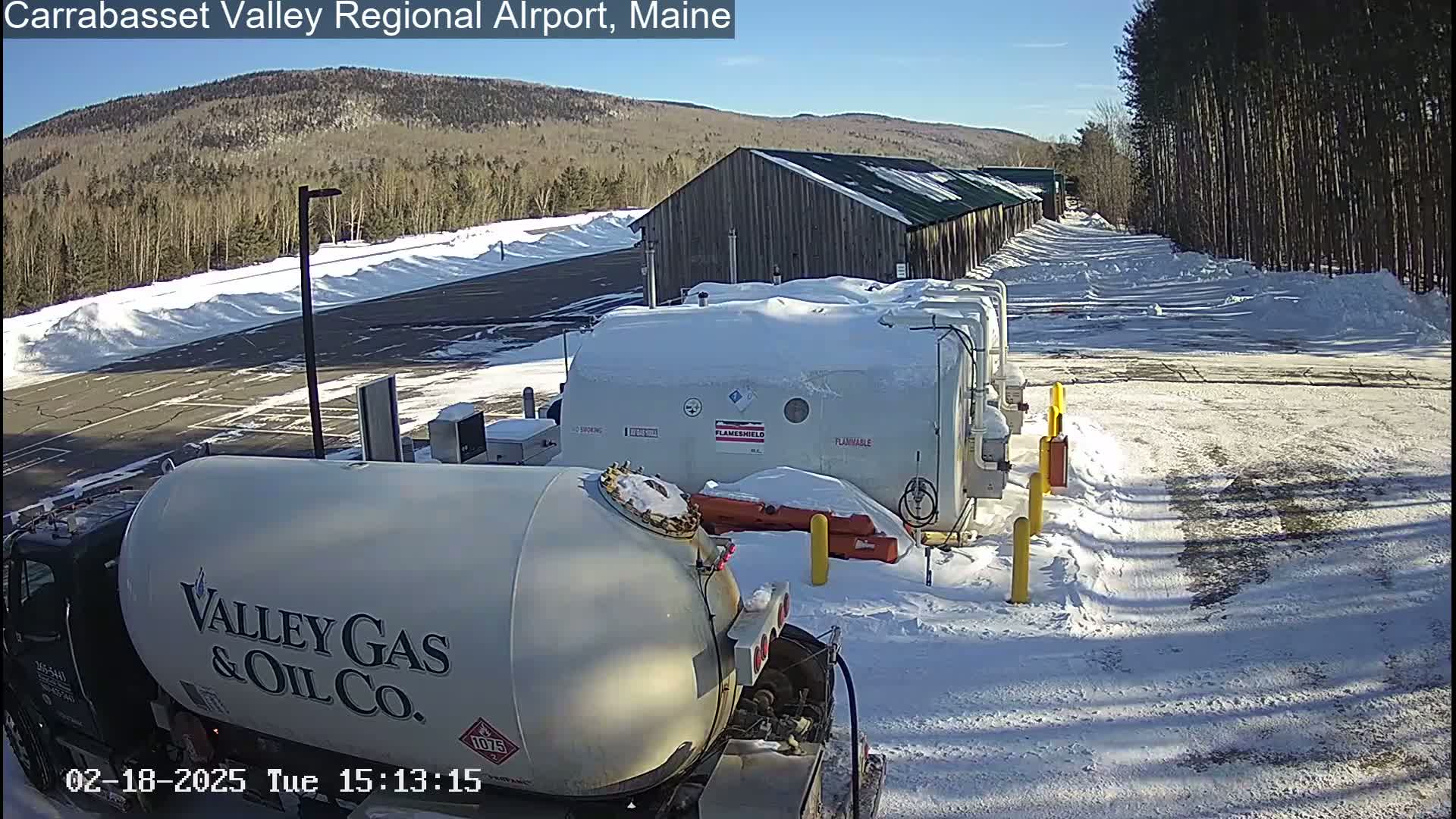 A snow-covered fuel depot with a gas truck in the foreground,  under a sunny winter sky, is situated next to a wood building and a line of trees.