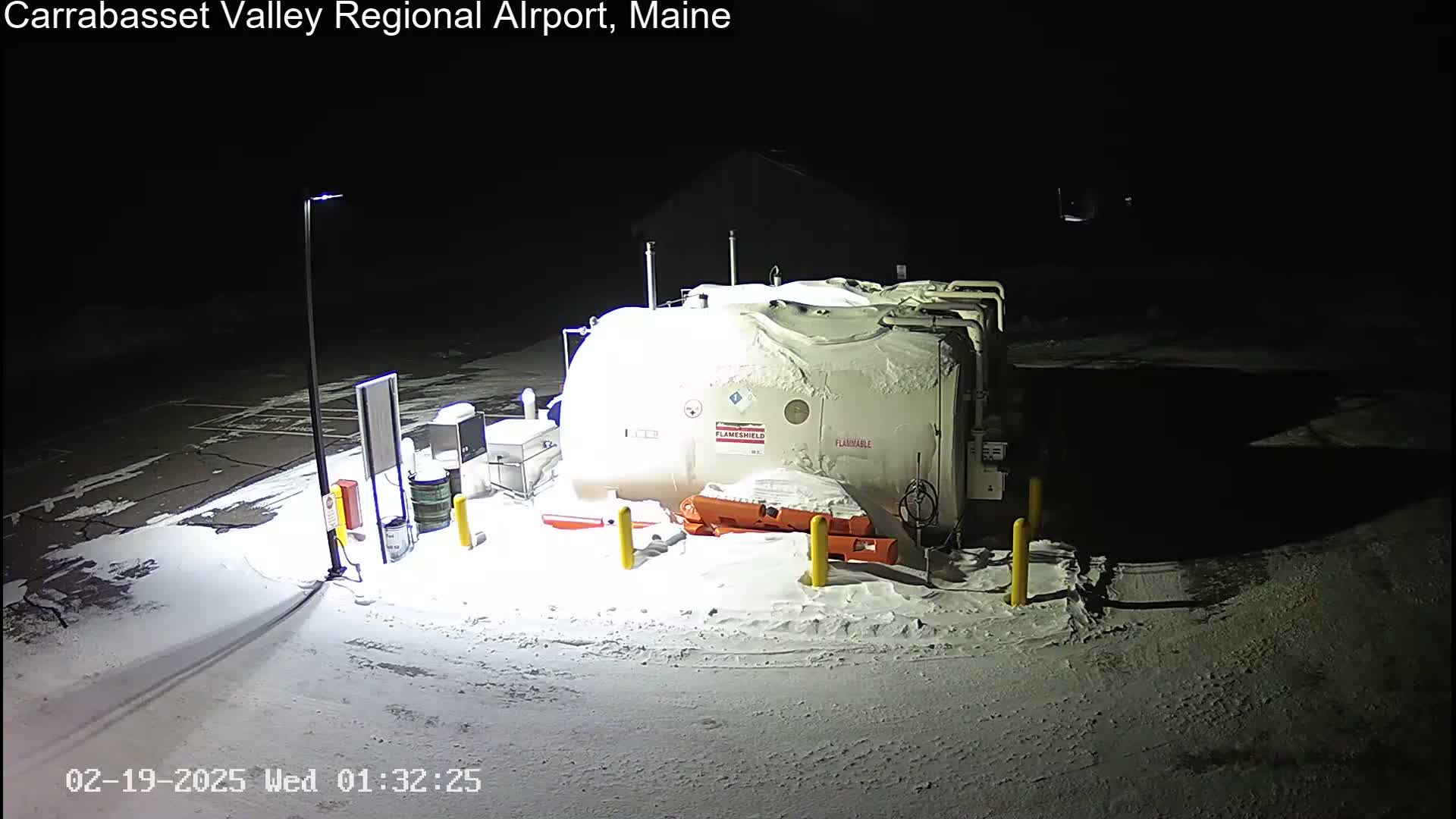 A large, snow-covered fuel tank sits in a snow-covered parking lot at night, illuminated by a single light pole.