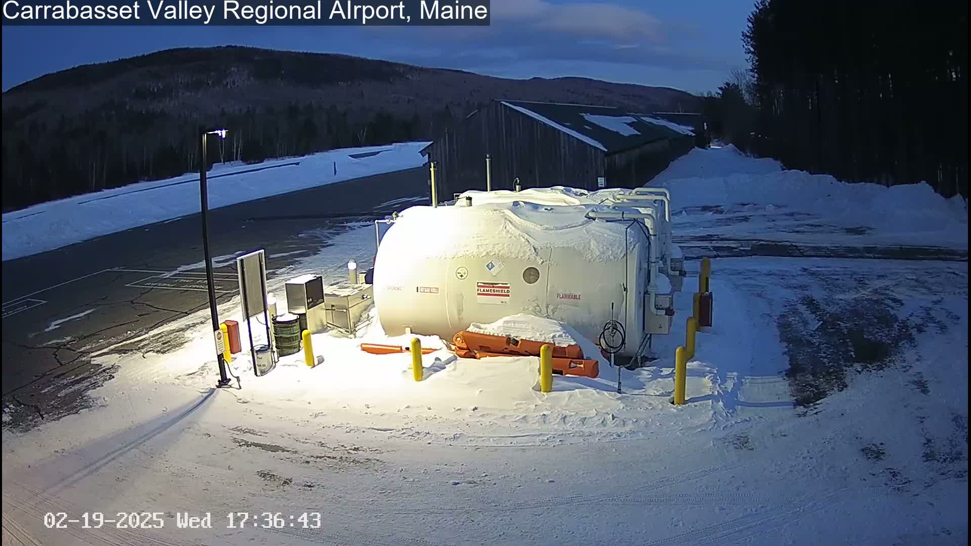 A large, snow-covered fuel tank sits in a snow-covered parking lot at dusk, with a building and a wooded hill visible in the background.