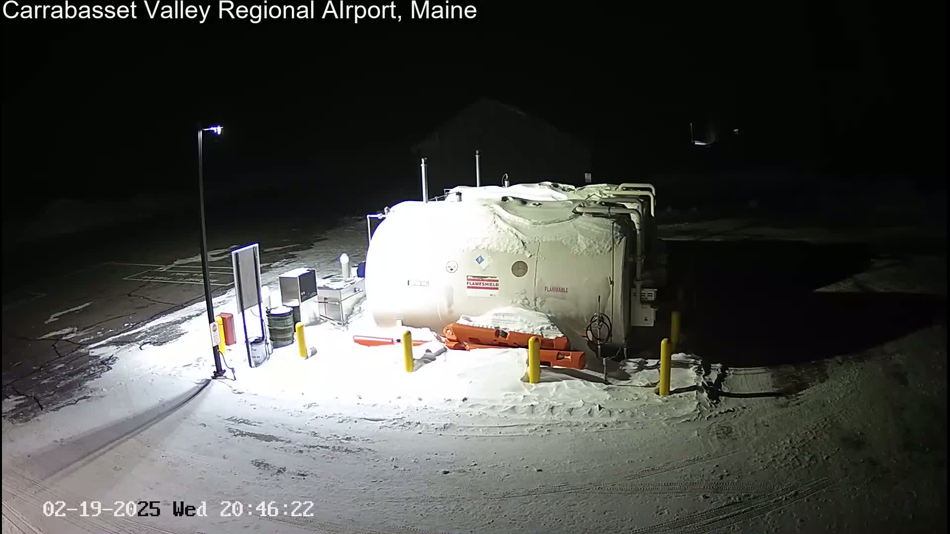 A large, snow-covered tank sits in a snow-covered parking lot at night, illuminated by a single lamppost.