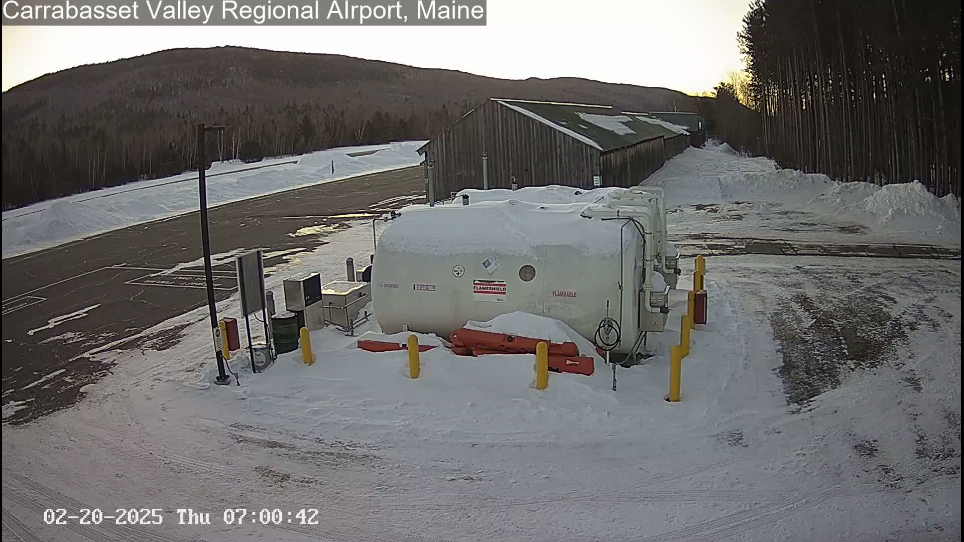 A snow-covered airport fueling station sits in a mostly empty parking lot next to a long wooden building, with a mountain range visible in the background under a clear, bright sky.