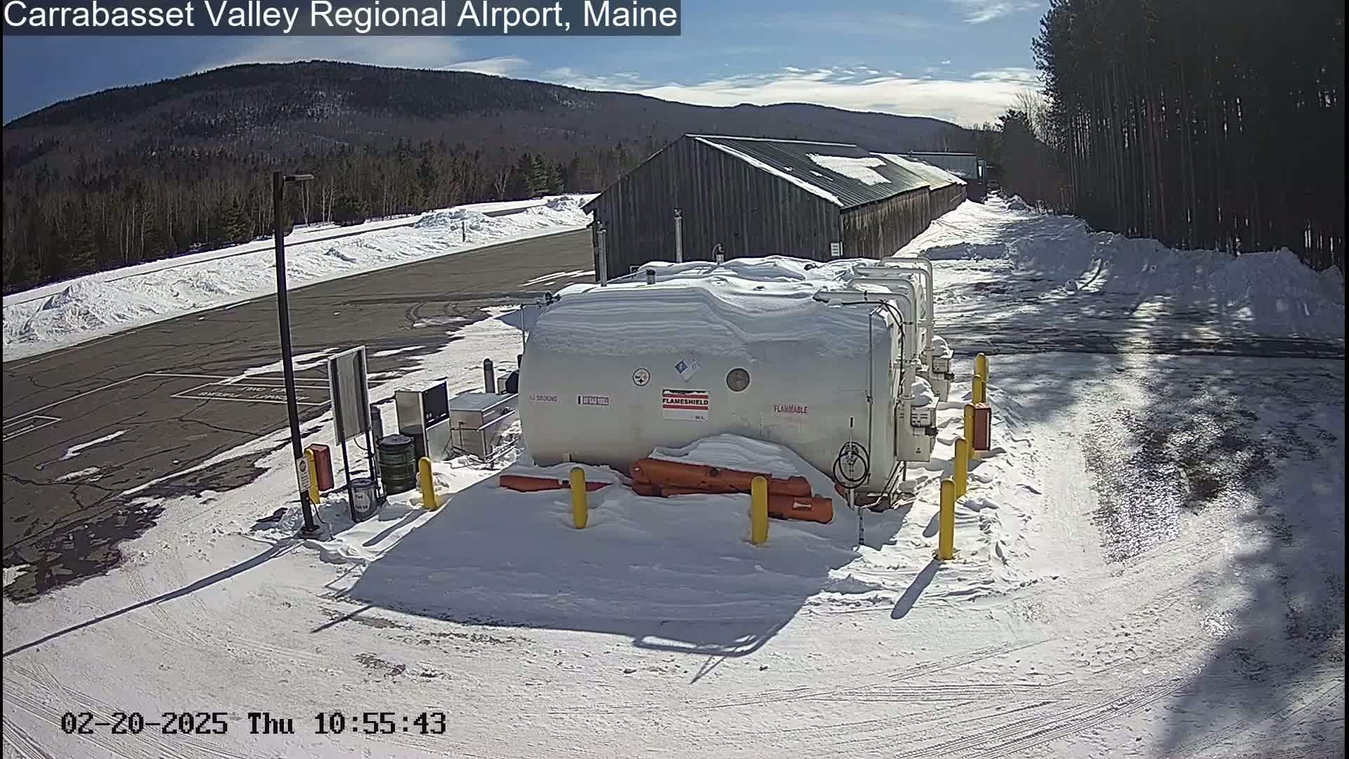 A snow-covered fuel storage area sits next to a long, gray building, with a sunny, snow-covered landscape and distant mountains visible in the background.