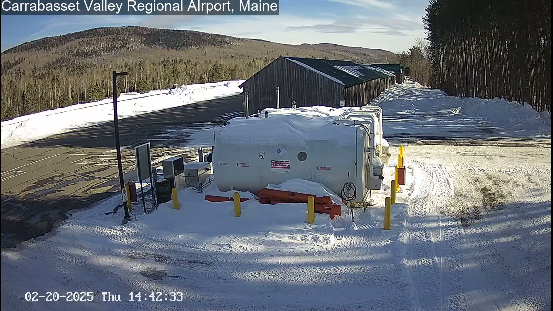 A snowy airport parking lot with a snow-covered fuel tank and a long wooden building in the background under a sunny sky.