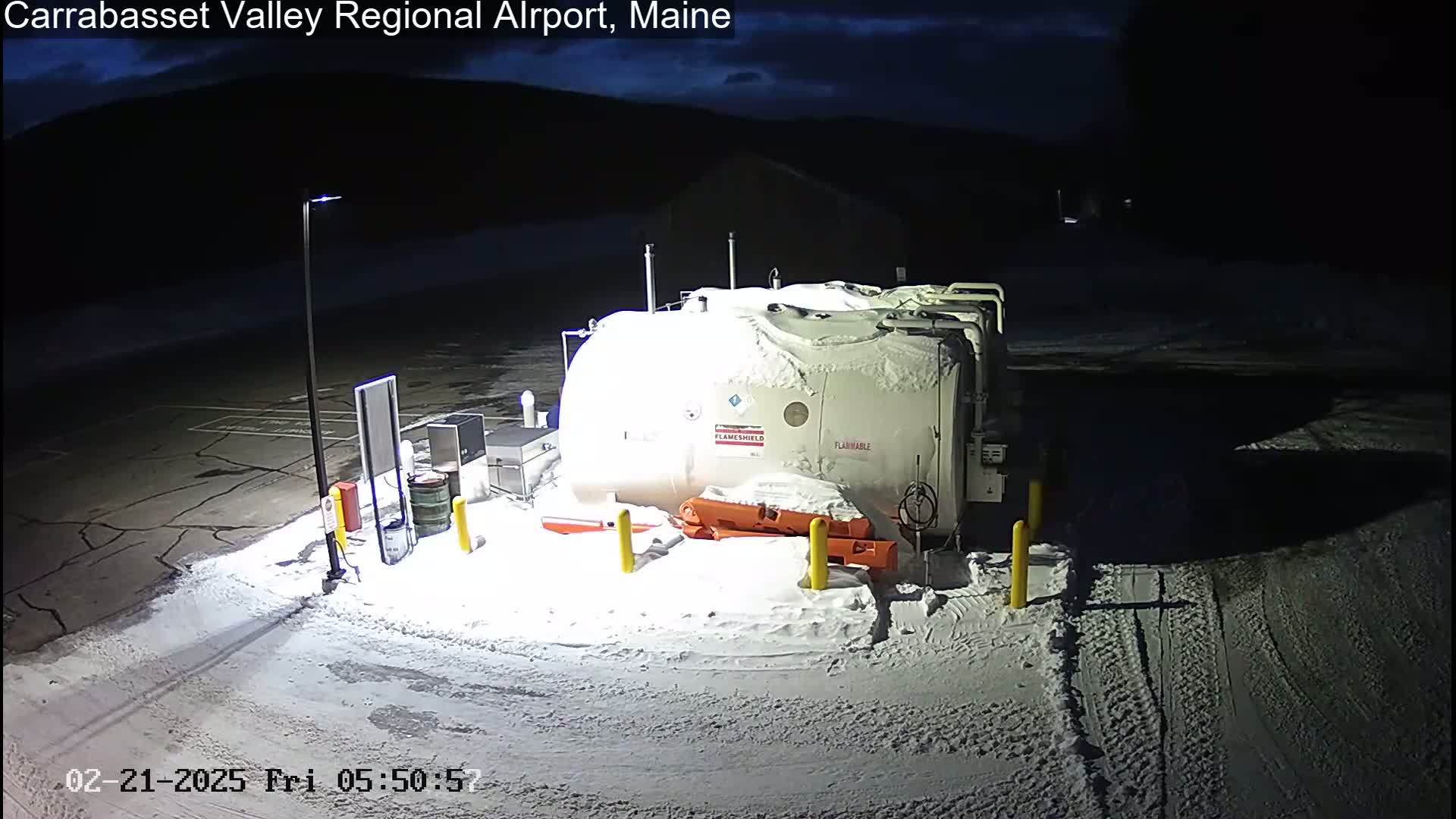 A snow-covered, large, white fuel tank sits in a mostly empty, snow-covered parking lot at night.