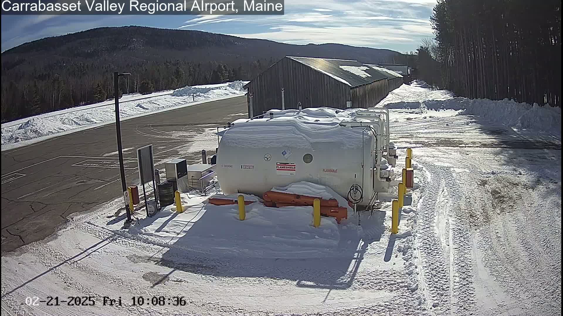 A snow-covered airport tarmac features several large, snow-covered fuel tanks near a long, wooden building under a sunny sky.