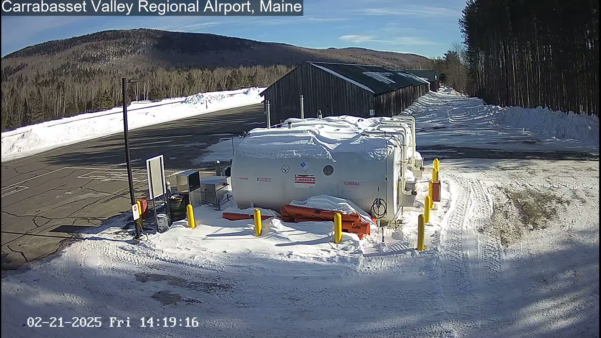 A snow-covered fuel storage area sits near a long, snow-covered building under a sunny sky, with mountains and trees visible in the background.