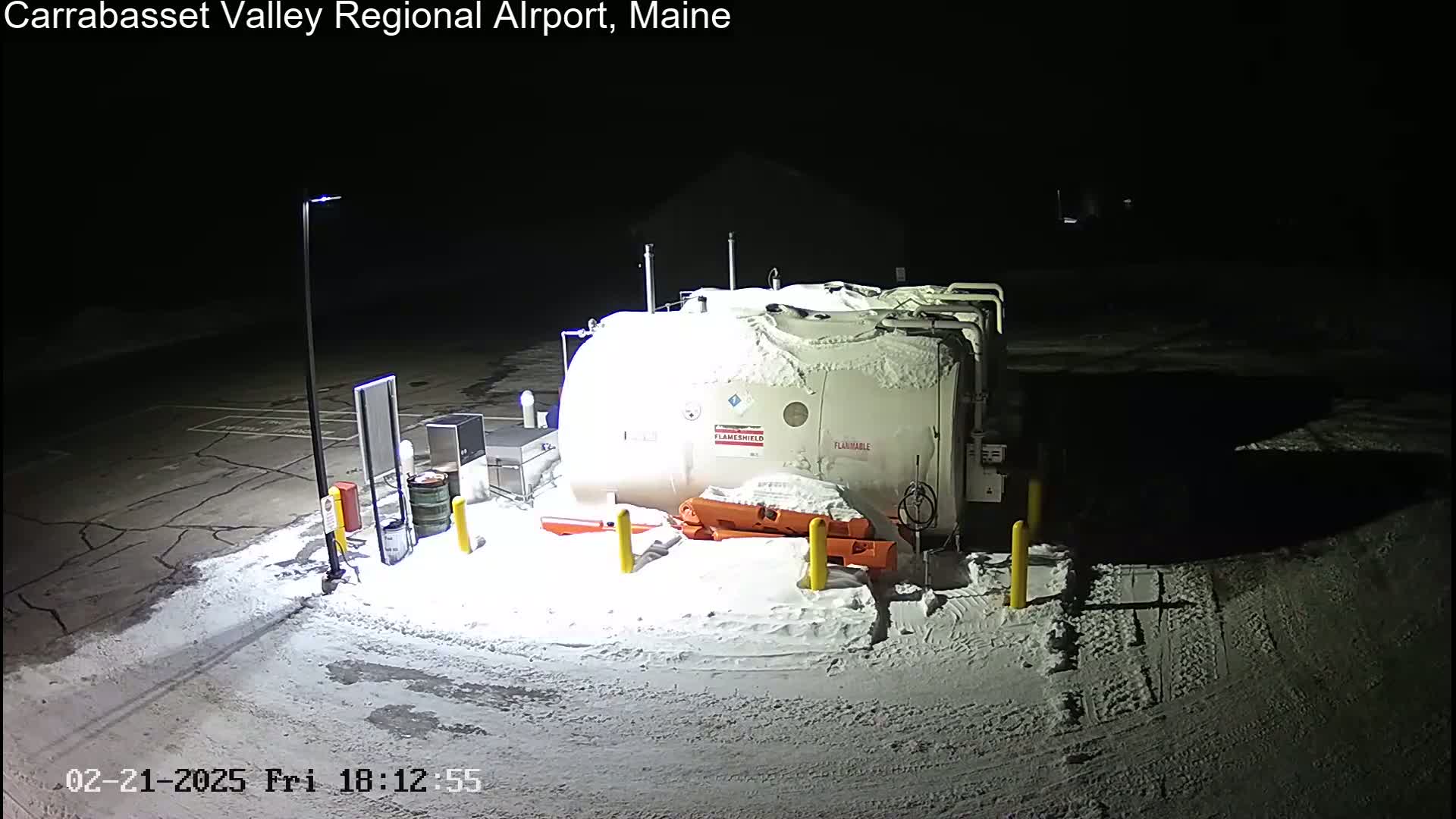 A large, snow-covered fuel tank sits in a snow-covered parking lot at night, illuminated by a single lamppost.