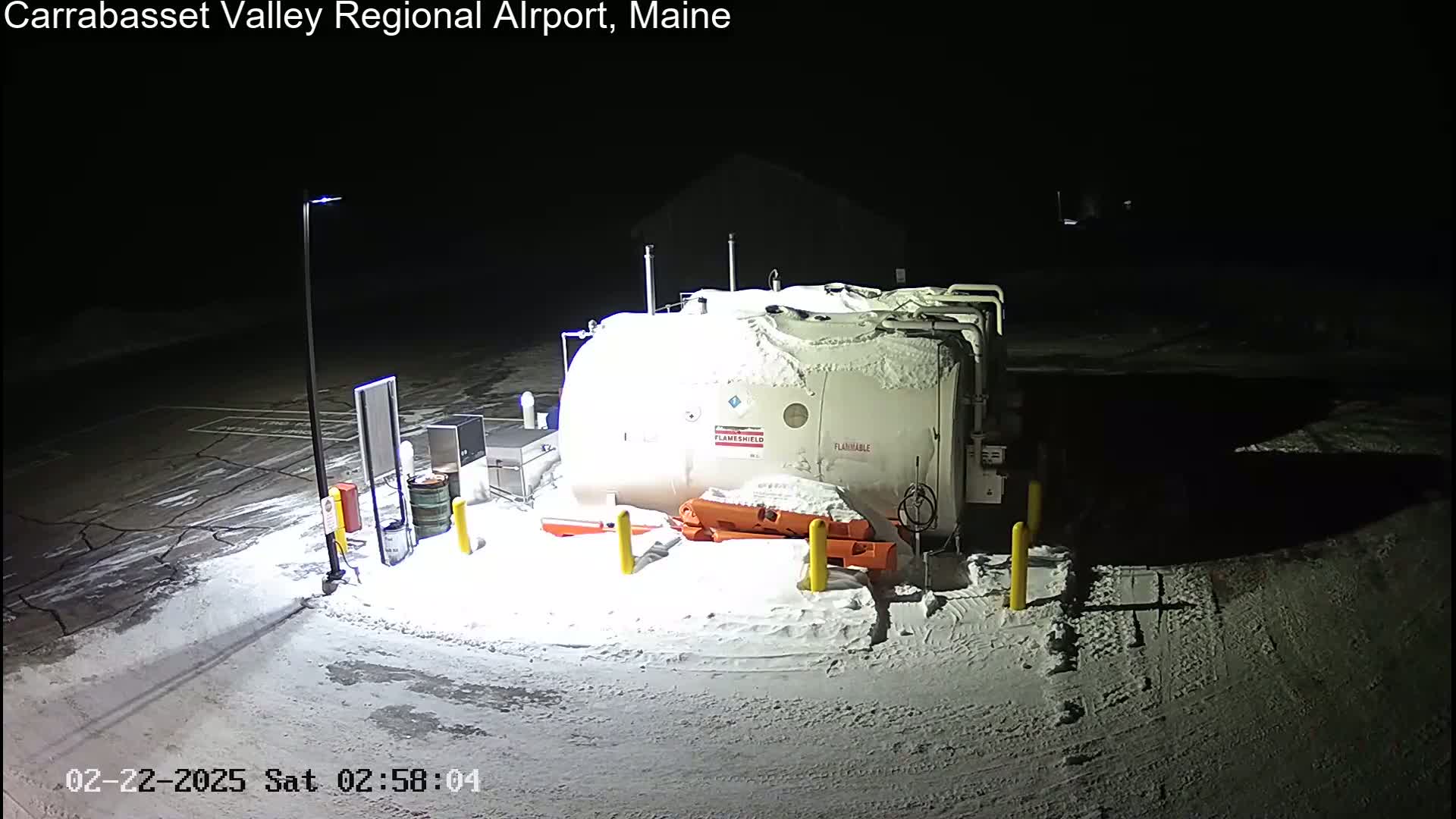 A large, snow-covered fuel tank sits in a snow-covered parking lot at night, illuminated by a single lamppost.