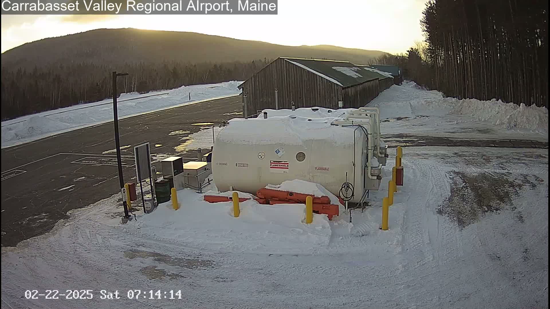 A snow-covered airport tarmac with a large white tank and a wooden building in the background, under a partly sunny sky.
