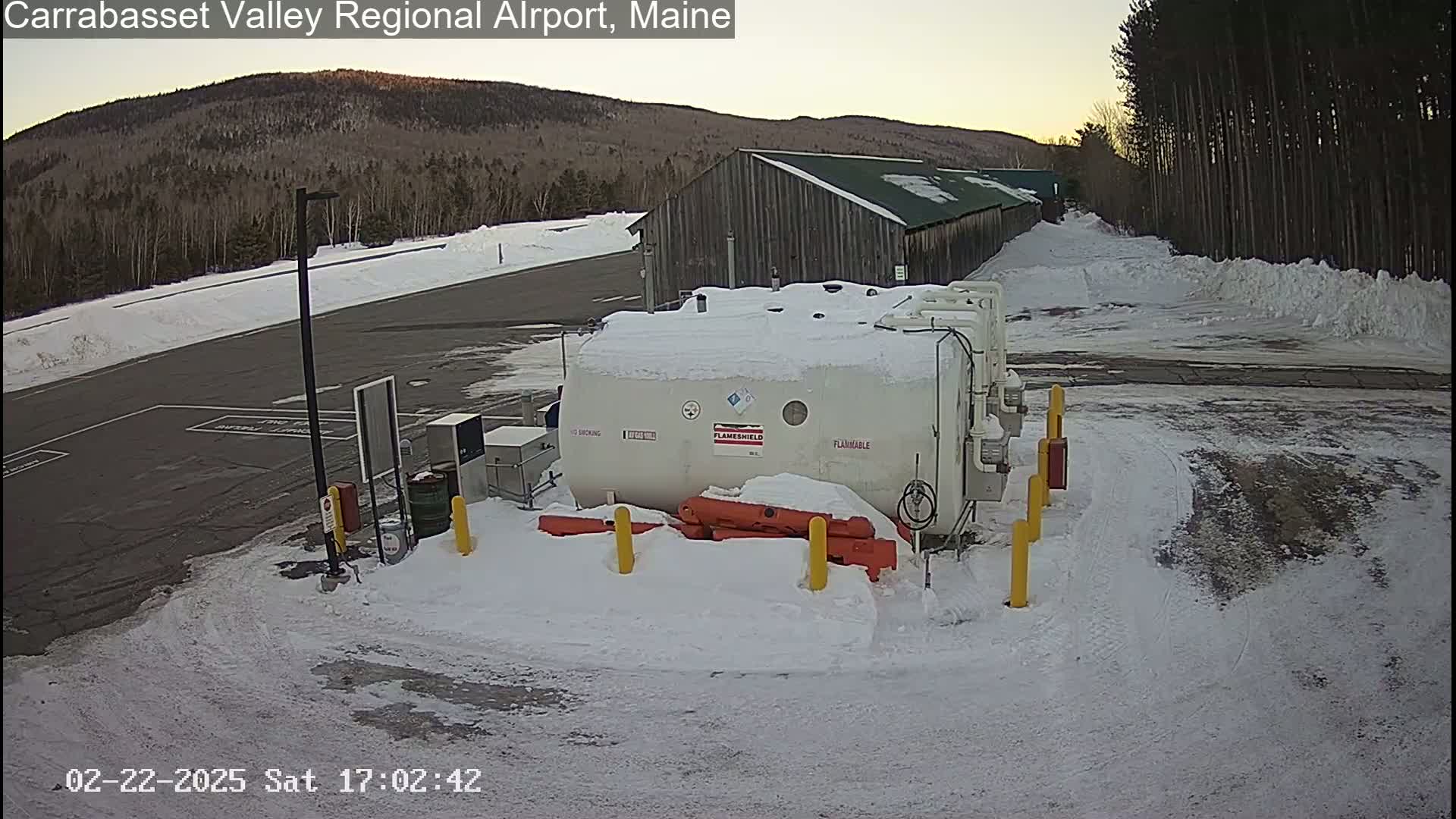 A snow-covered airport tarmac with a large, white, flammable tank, a long building, and a backdrop of a snow-dusted mountain range under a twilight sky.