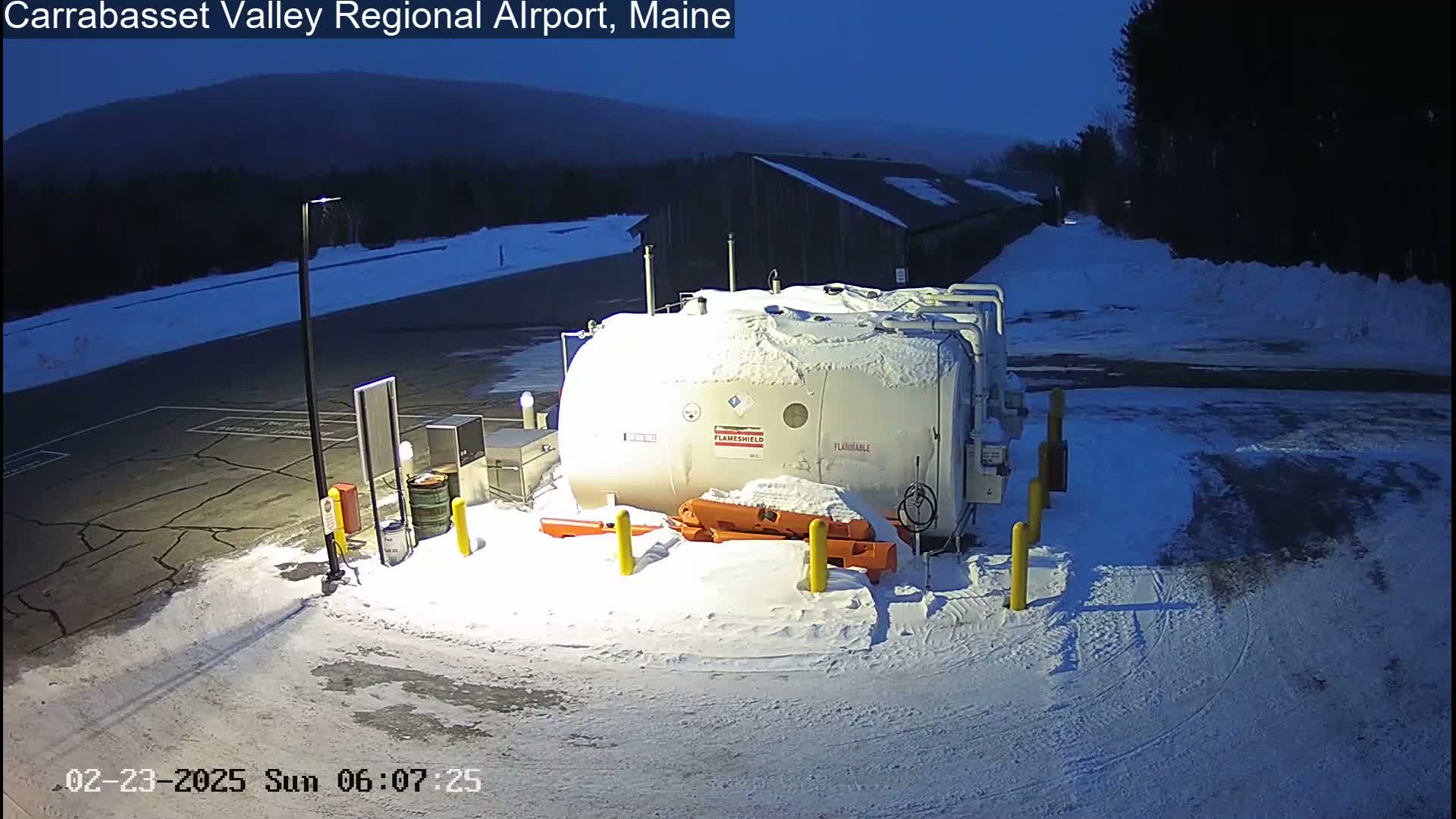 A large, snow-covered fuel tank sits in a snow-covered parking lot at dusk, near a dark building and some trees.